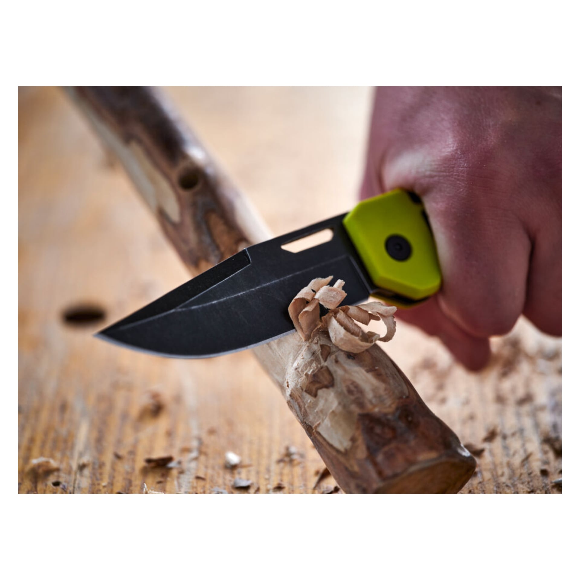 En hand använder den grönskaftade Ryobi-jackkniven RHCKF-3 för att skära tunna träremsor från en liten stock som ligger på en träyta.