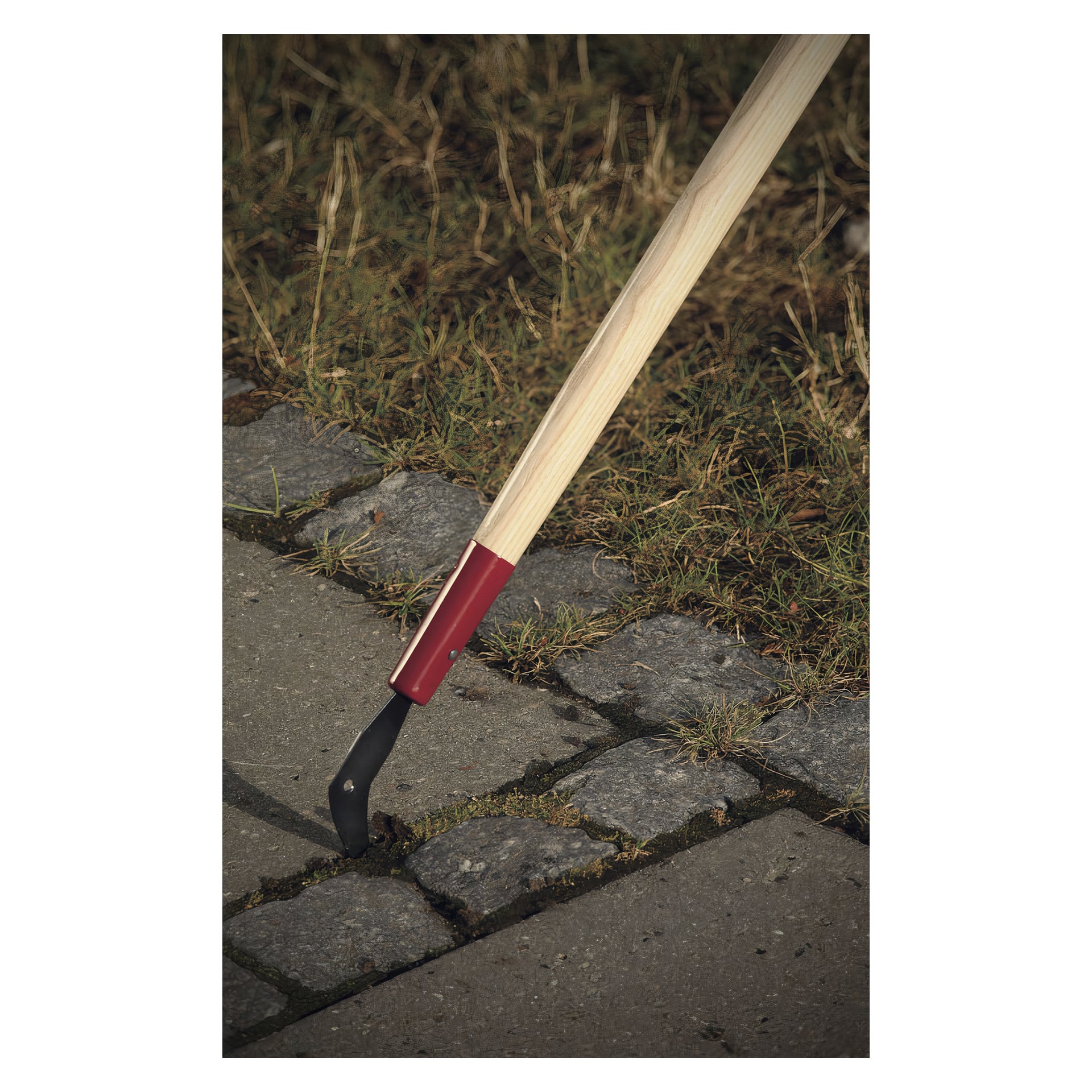 En Fiskars patio-skrapa med långt trähandtag och svart metallblad är designad för att rensa ogräs från springorna mellan betong- och stenplattor som kantas av gräs och jord.