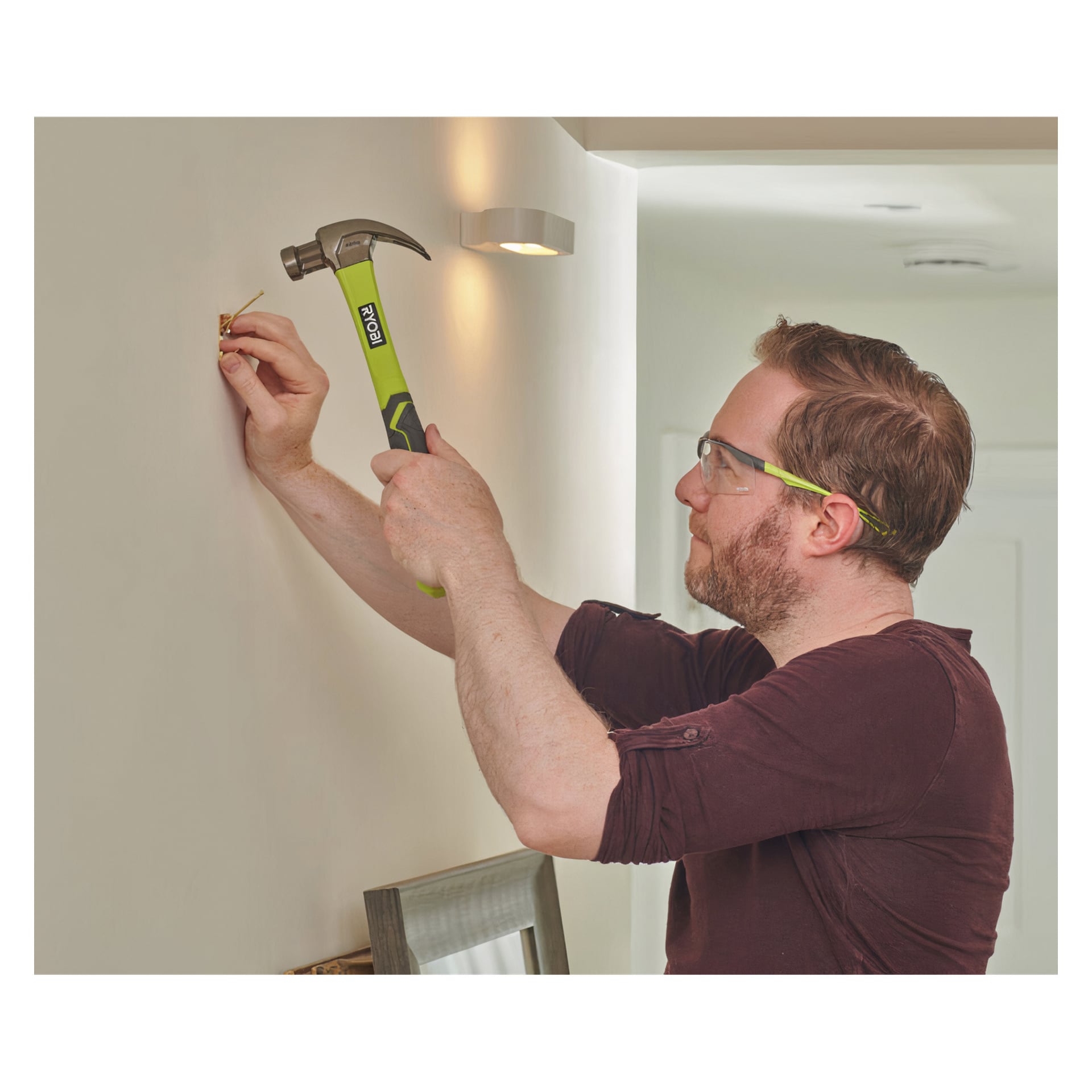 En man med skyddsglasögon använder en grön Ryobi Carpenter's Hammer RHHCC450 för att installera ett fäste i en inomhusvägg med vit. I närheten syns en lampa och en del av en träram.