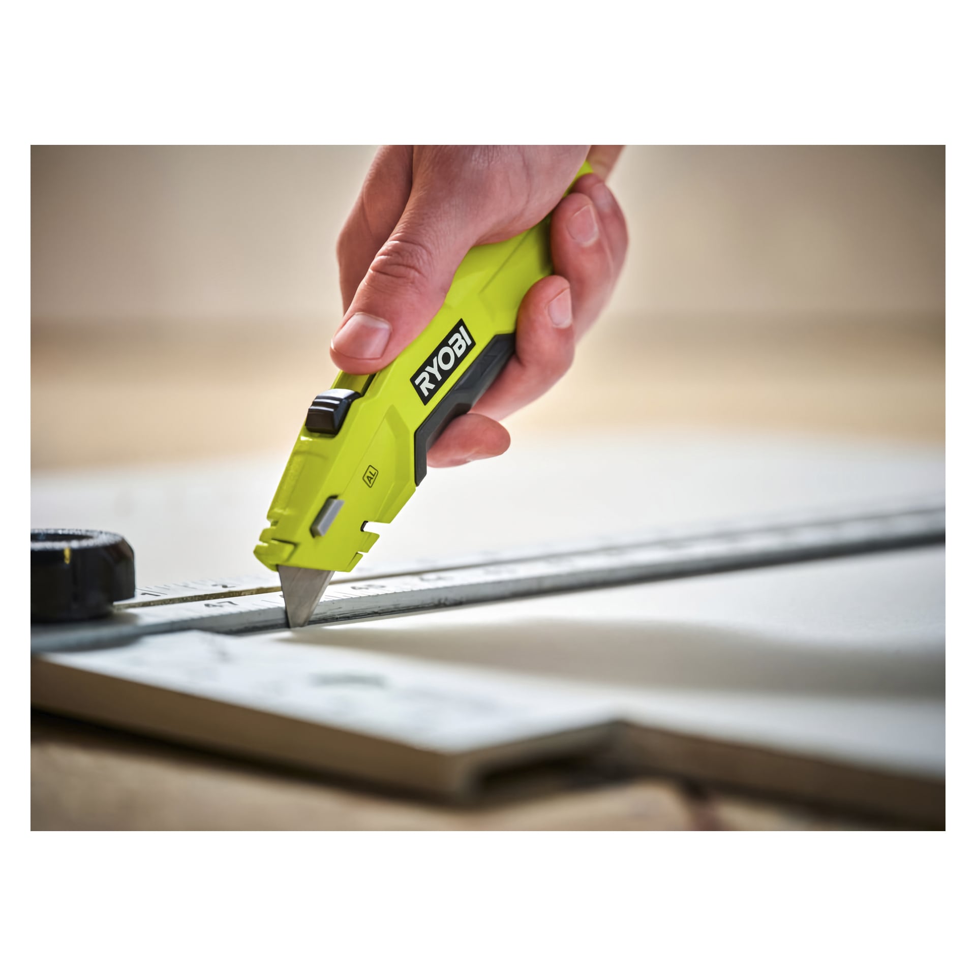 En hand håller en grön Ryobi Universal-kniv RHCKF-1 och skär material längs kanten på en metalllinjal; Ett måttband vilar på vänstra sidan av arbetsytan.