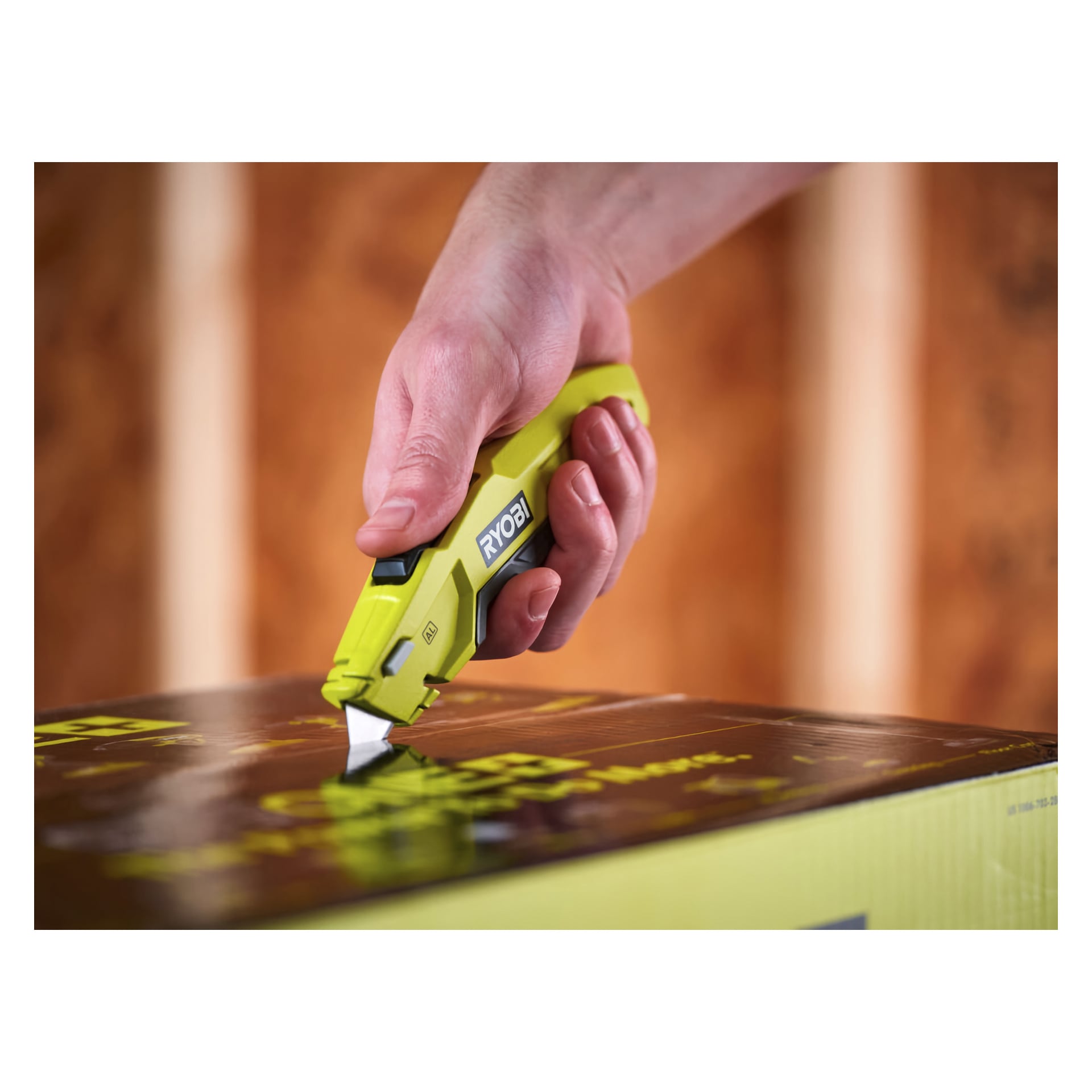 En grön Ryobi Universal-kniv RHCKF-1 hålls i handen och skär igenom tejp som förseglar en kartonglåda. Den suddiga bakgrunden har vertikala träbjälkar.