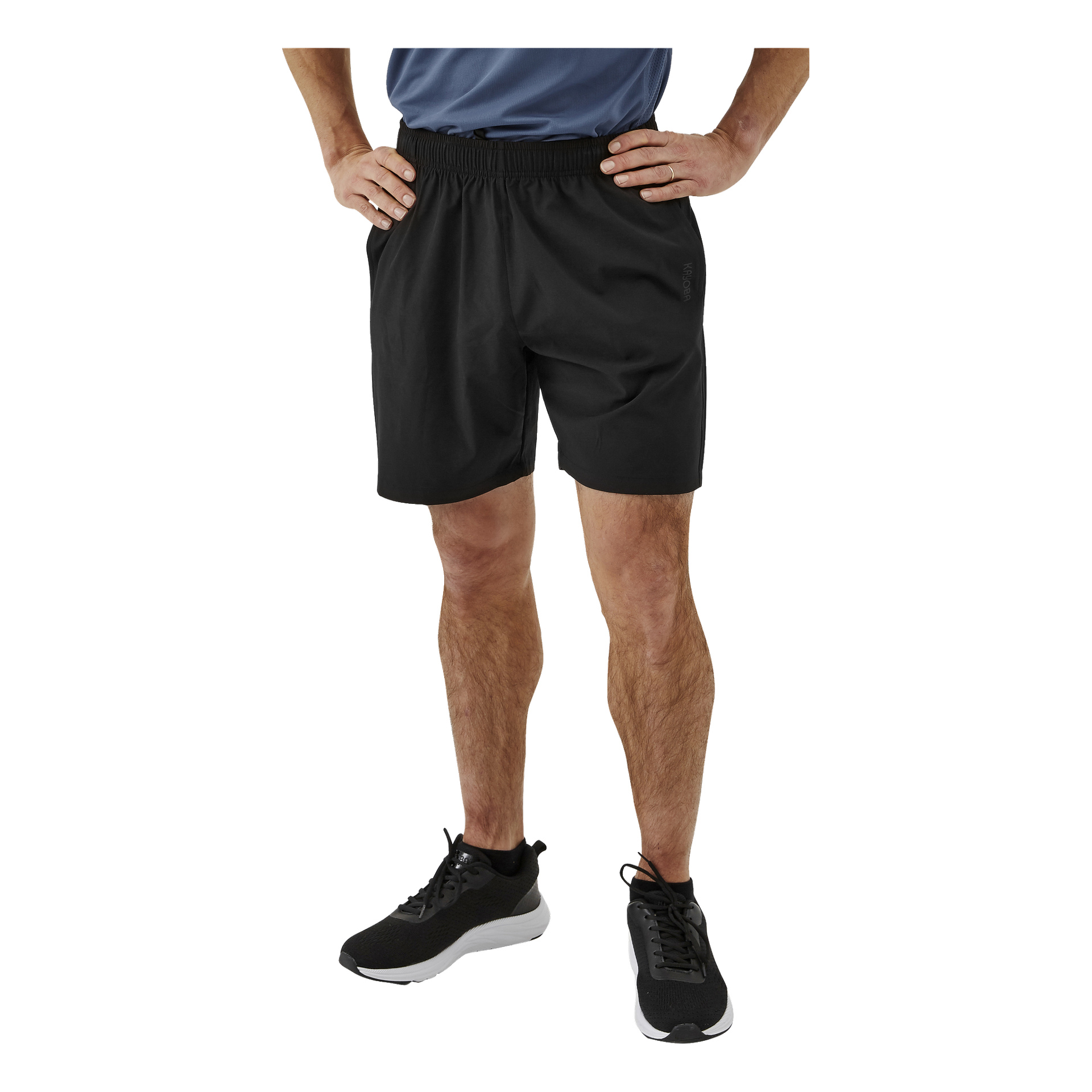 En person är synlig från midjan och neråt, stående med händerna på höfterna. De bär svarta Kayoba herrträningsshorts, en blå tröja och svarta löparskor med vita sulor.