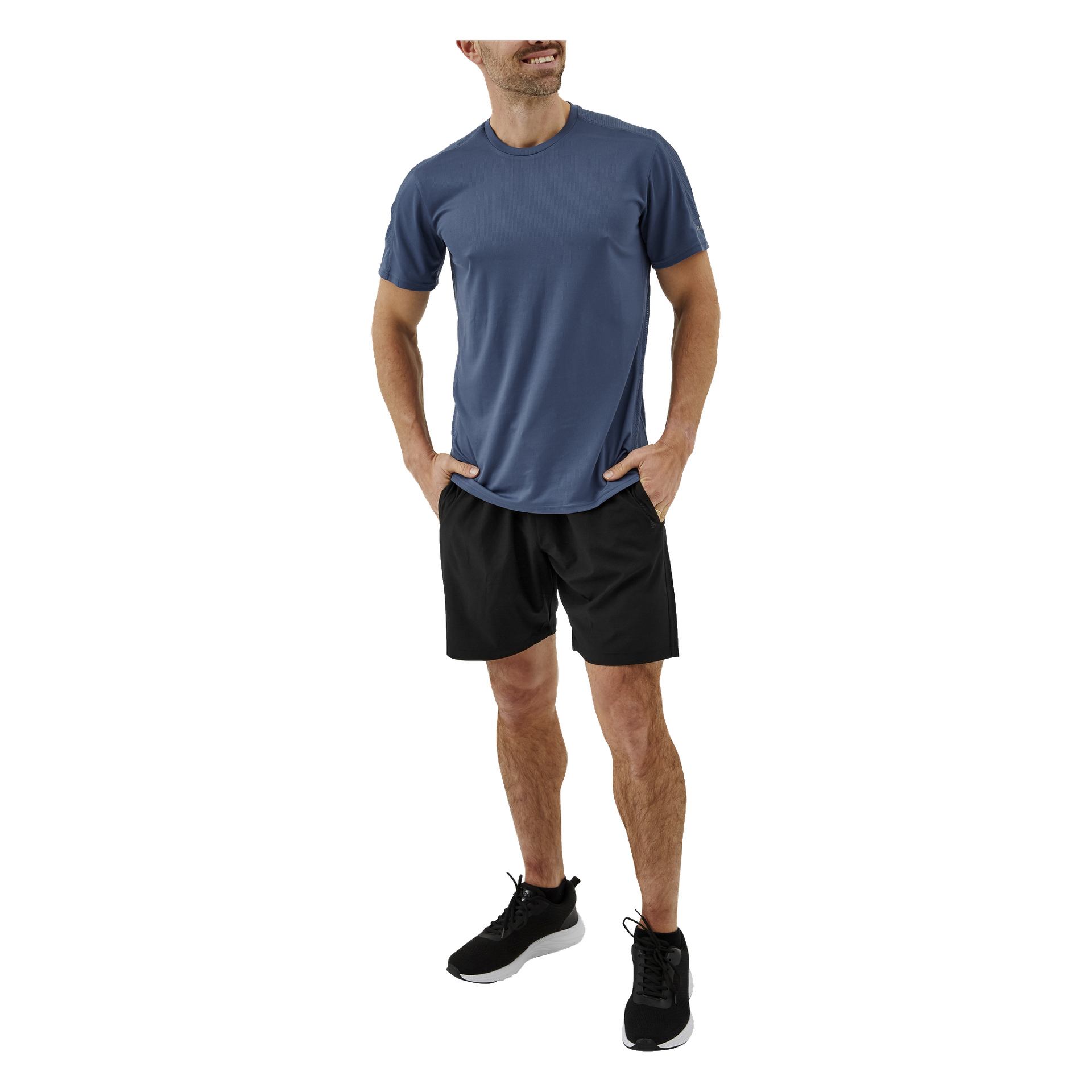 En man med kort skägg står med händerna i fickorna, klädd i Kayoba herrträningsshorts, en blå kortärmad T-shirt och svarta träningsskor. Han ler och tittar lite åt vänster.