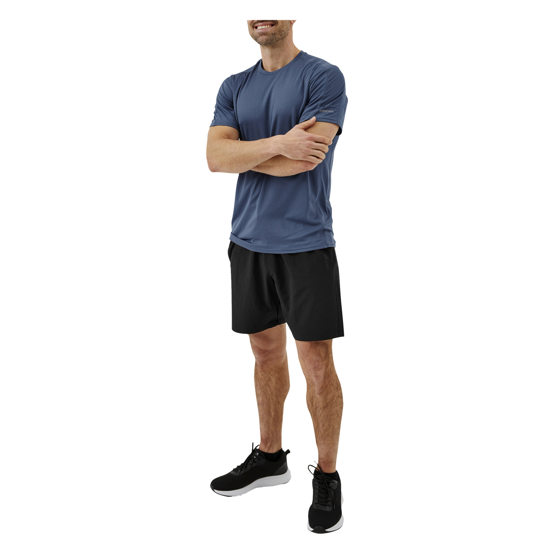 En leende man står med armarna i kors, klädd i Kayoba Men's träningsshorts, en blå kortärmad tröja och svarta löparskor med vita sulor. Bilden är beskuren precis ovanför hans huvud.