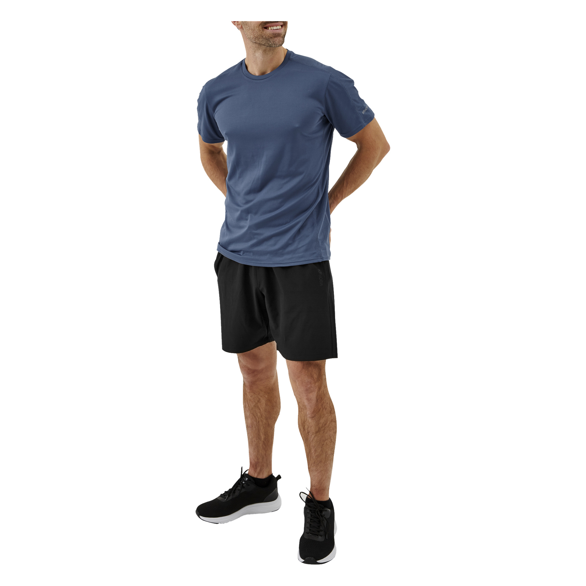 En man står i en avslappnad pose med händerna bakom ryggen, iklädd Kayoba Men's träningsshorts, en blå T-shirt och svarta träningsskor. Han ler och tittar åt vänster.