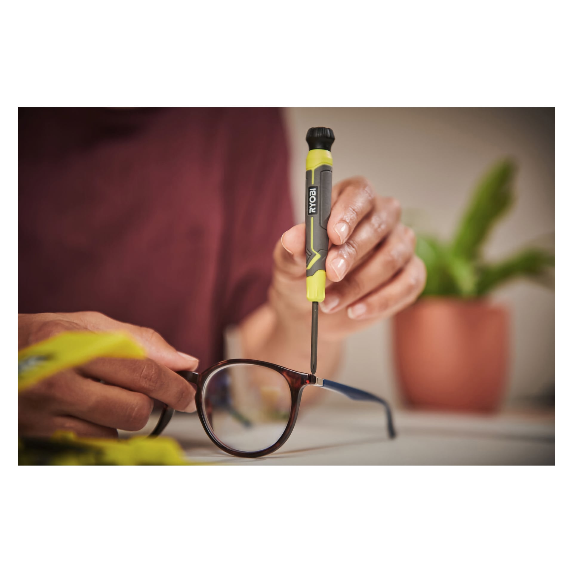 En person använder en gul och svart Ryobi Precision Screwdriver från RHSDSPC6 sexdelade set för att dra åt en gångjärnsskruv på glasögon, med en suddig orange krukväxt i bakgrunden.