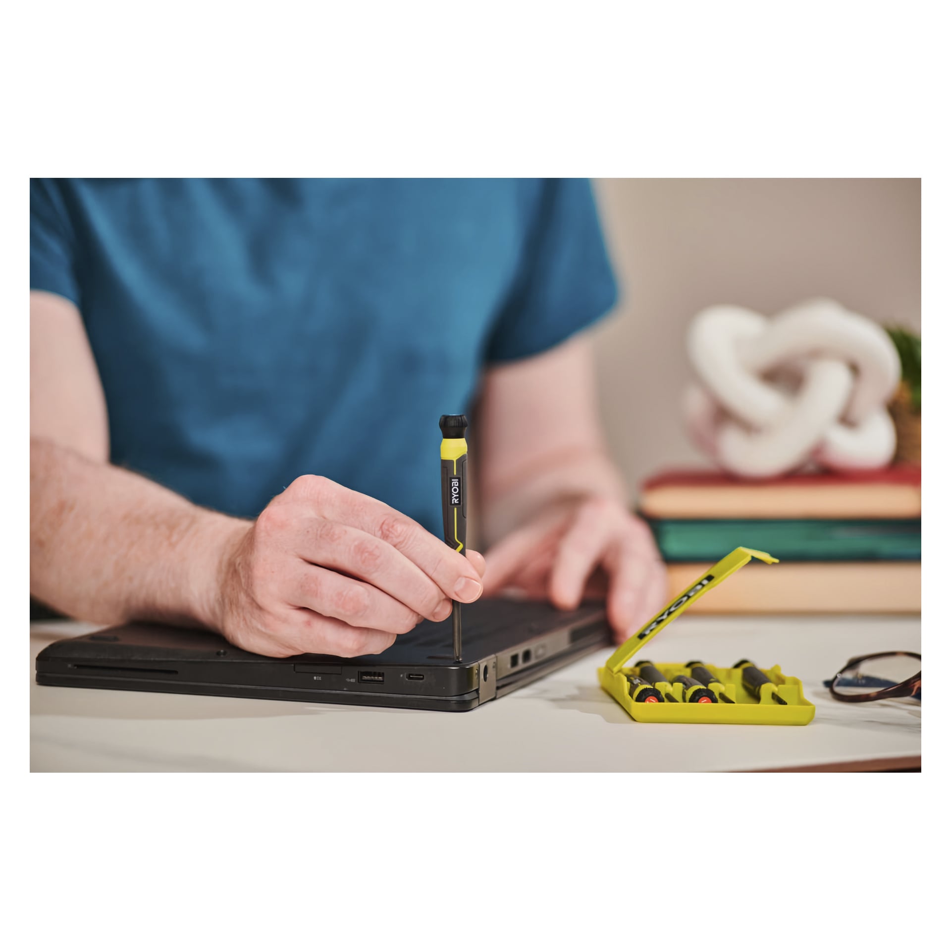 Någon arbetar på en svart laptop med en gul och svart Ryobi Precision Screwdriver Set RHSDSPC6. Det öppna sexdelade verktygslådan visar flera skruvmejslar i närheten, med suddiga böcker och ett dekorativt föremål i bakgrunden.