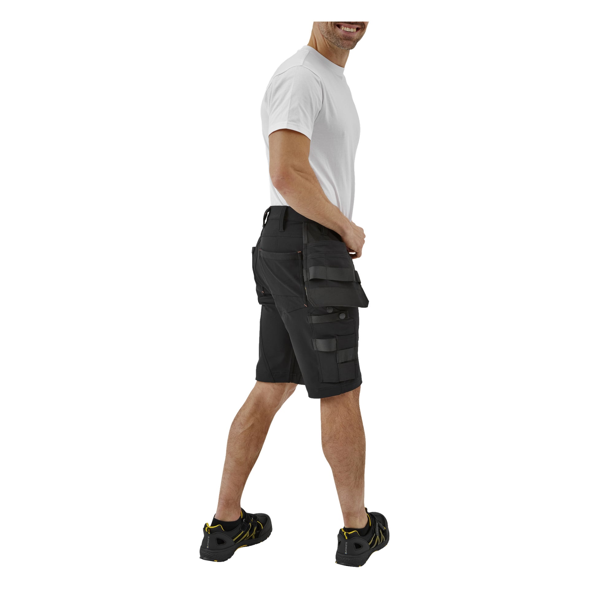 En leende man står lätt vinklad, klädd i en vit t-shirt, Bluewear Pro Workwear Steve herrshorts med flera fickor och svarta sportskor med gula detaljer.