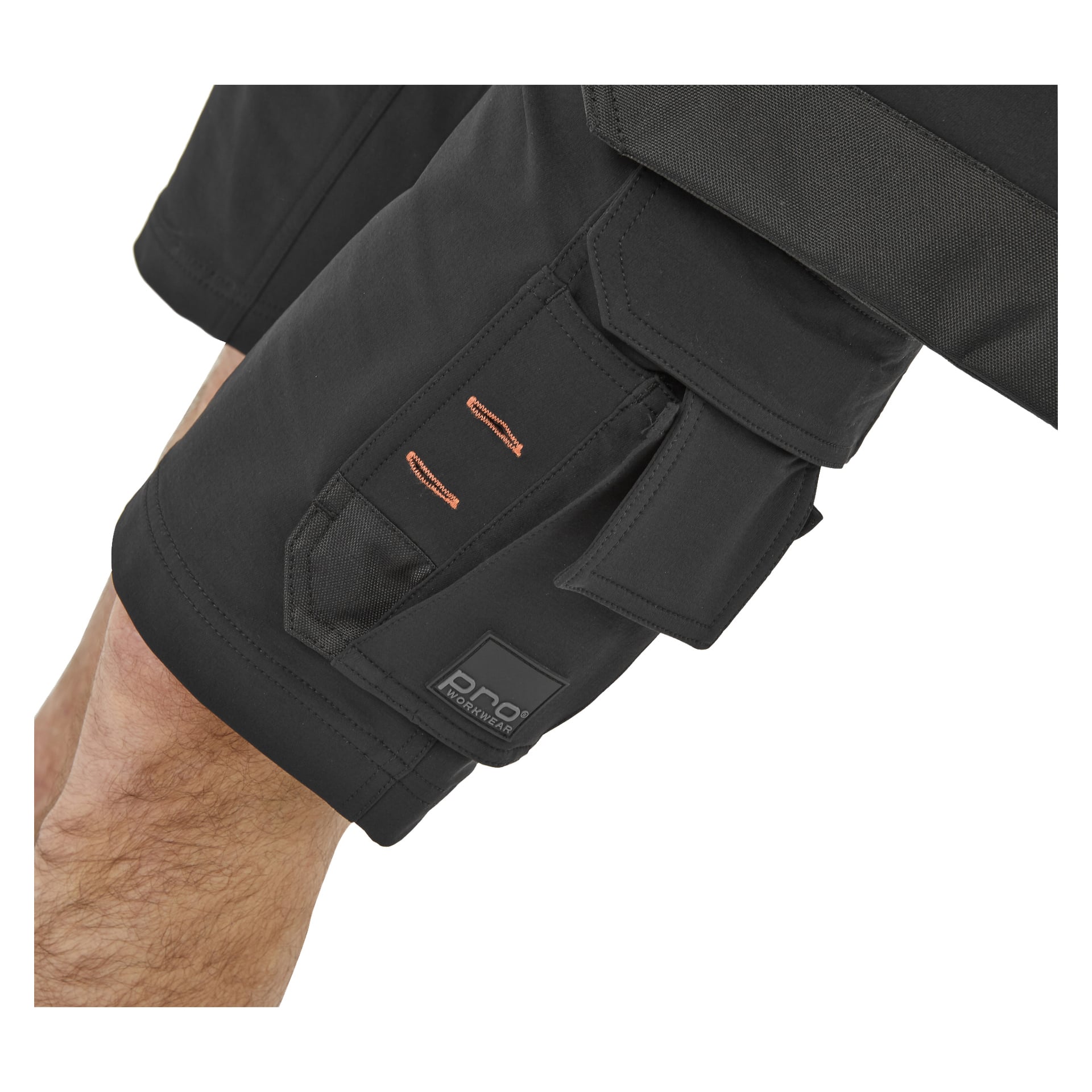 Närbild på en person klädd i Bluewear Pro Workwear svarta herrshorts Steve, med en sidoficka accentuerad med orange sömmar och en liten rektangulär etikett med "PRO." Endast en del av personens ben och shorts kan ses.