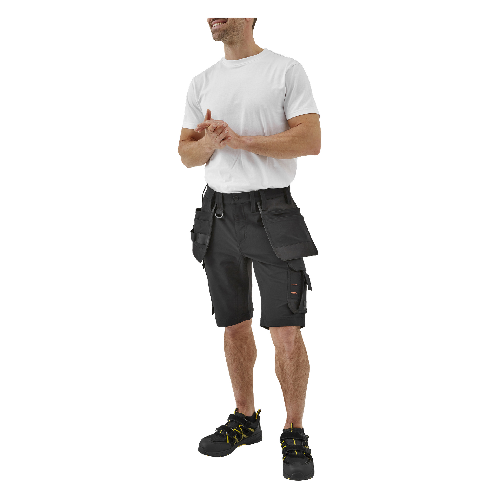 En leende man bär en enkel vit T-shirt, Bluewear Pro Workwear Steve-shorts med flera fickor och svarta arbetsskor med gula detaljer. Hans händer är knäppta framför bröstet.