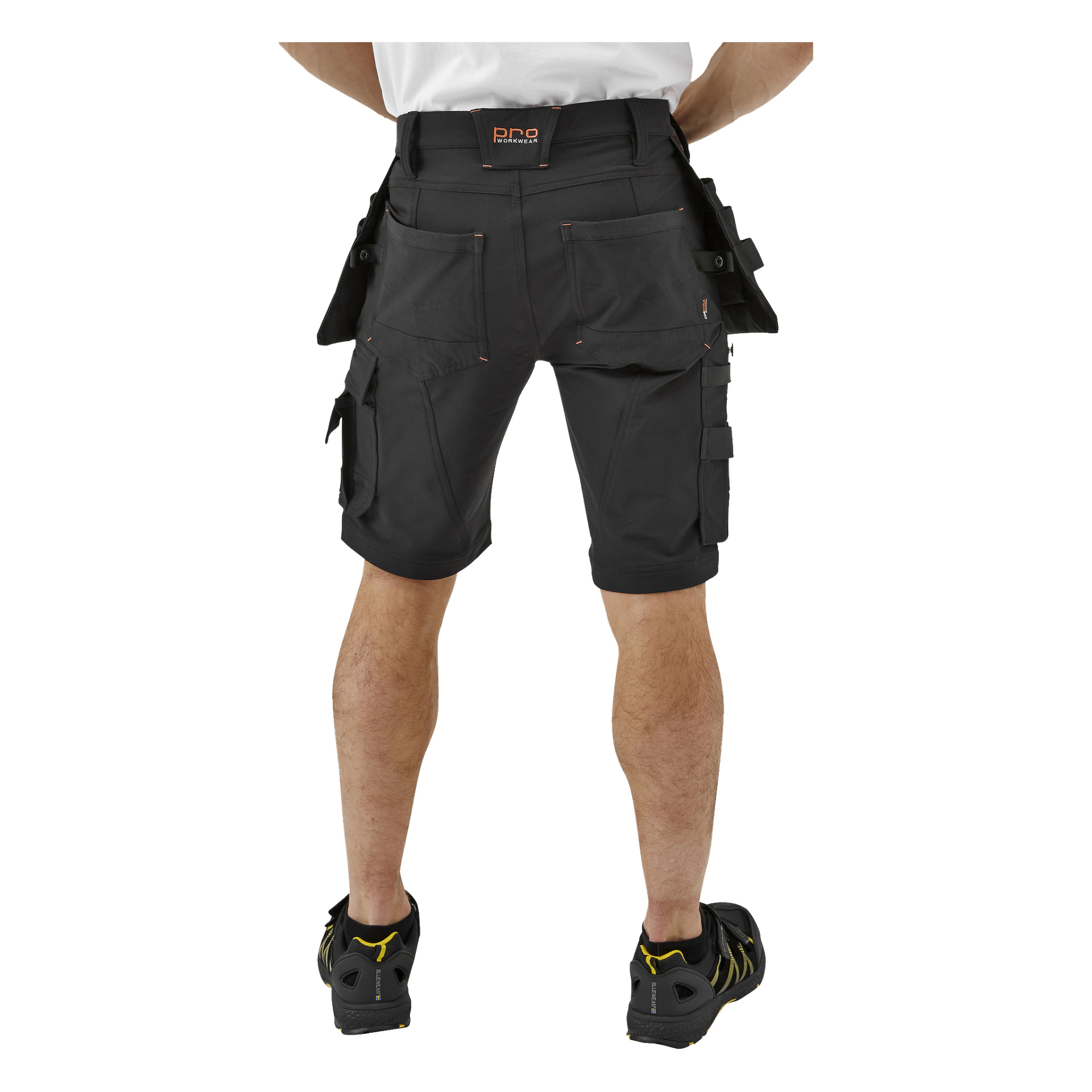 En person syns bakifrån, klädd i en vit skjorta, svarta Bluewear Pro Workwear Steve herrshorts med flera fickor och svarta sportskor med gula detaljer.