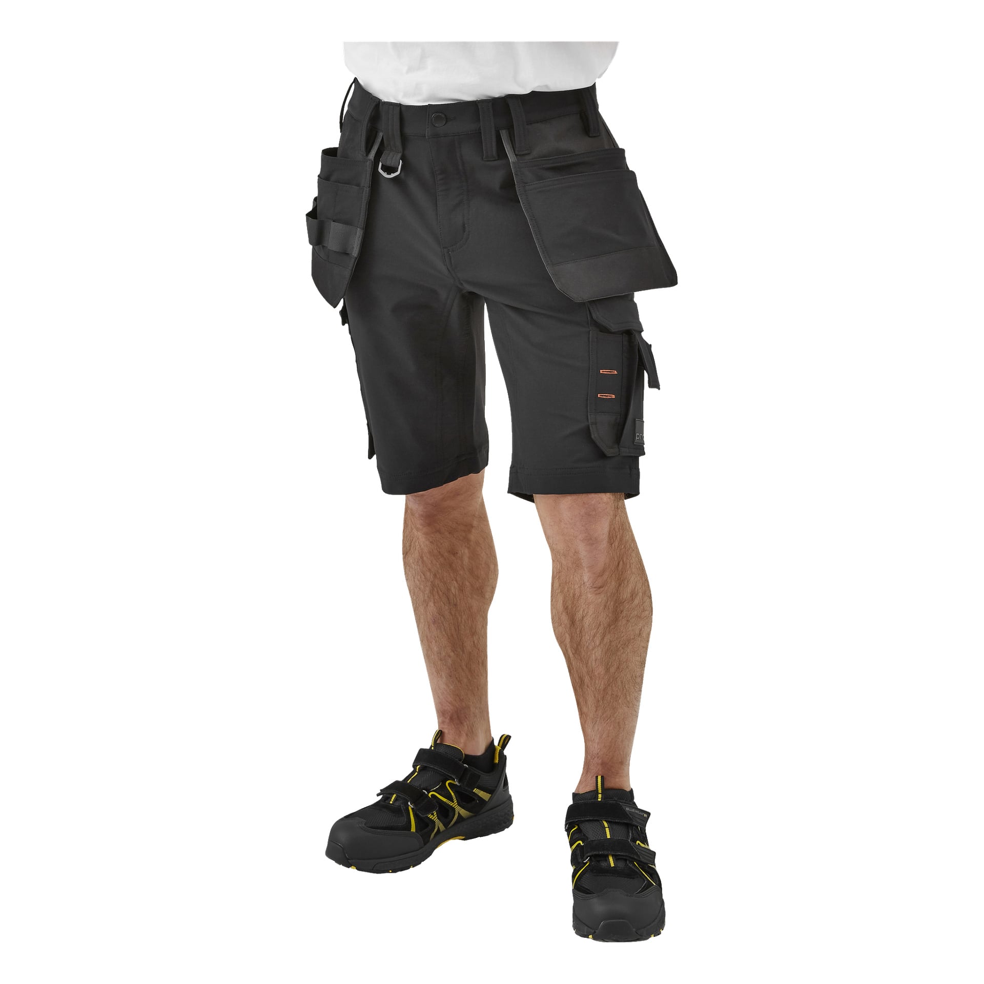 En vy från midjan och nedåt visar någon klädd i Bluewear Pro Workwear Steve herrshorts för arbete—svarta med flera fickor och verktygsfickor—matchade med svarta arbetsskor med gula detaljer. Bilden fokuserar på kläderna och utelämnar andra omgivningar.