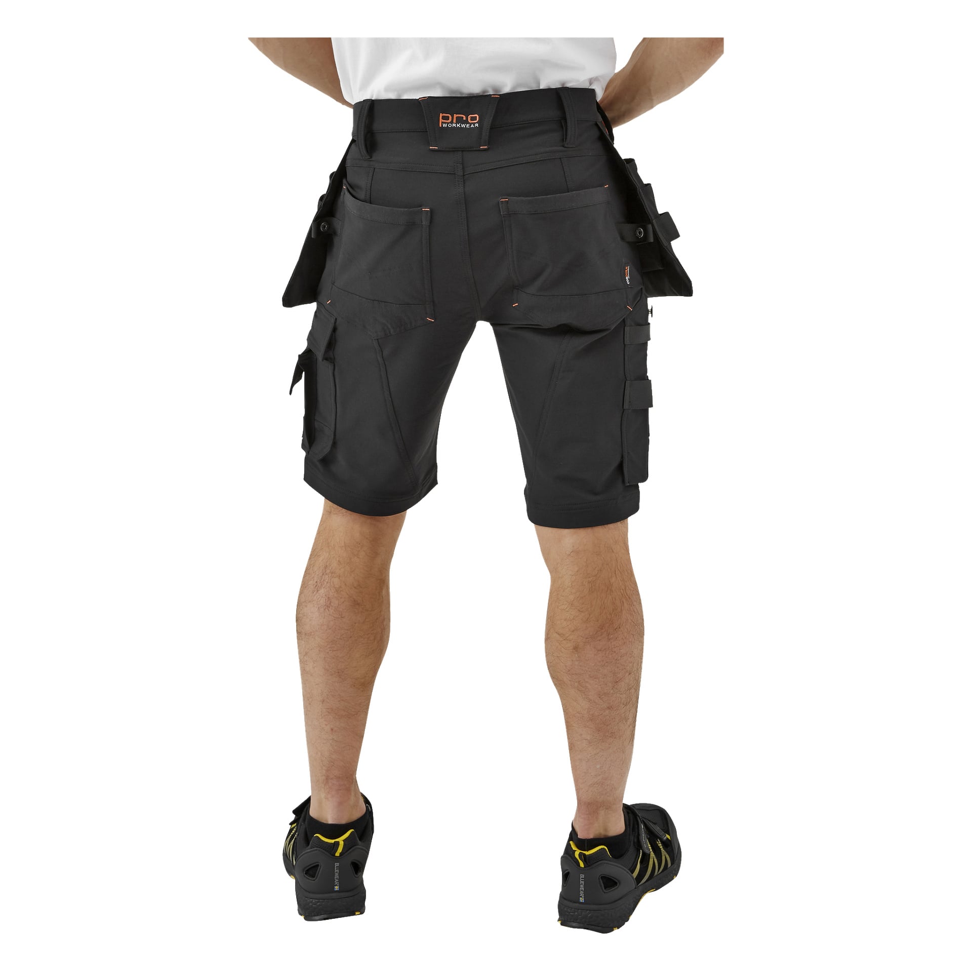En person syns bakifrån, iklädd svarta Bluewear Pro Workwear Men's Steve-shorts med flera fickor. De har på sig en vit skjorta instoppad i shortsen och svarta sneakers, stående med händerna på höfterna.