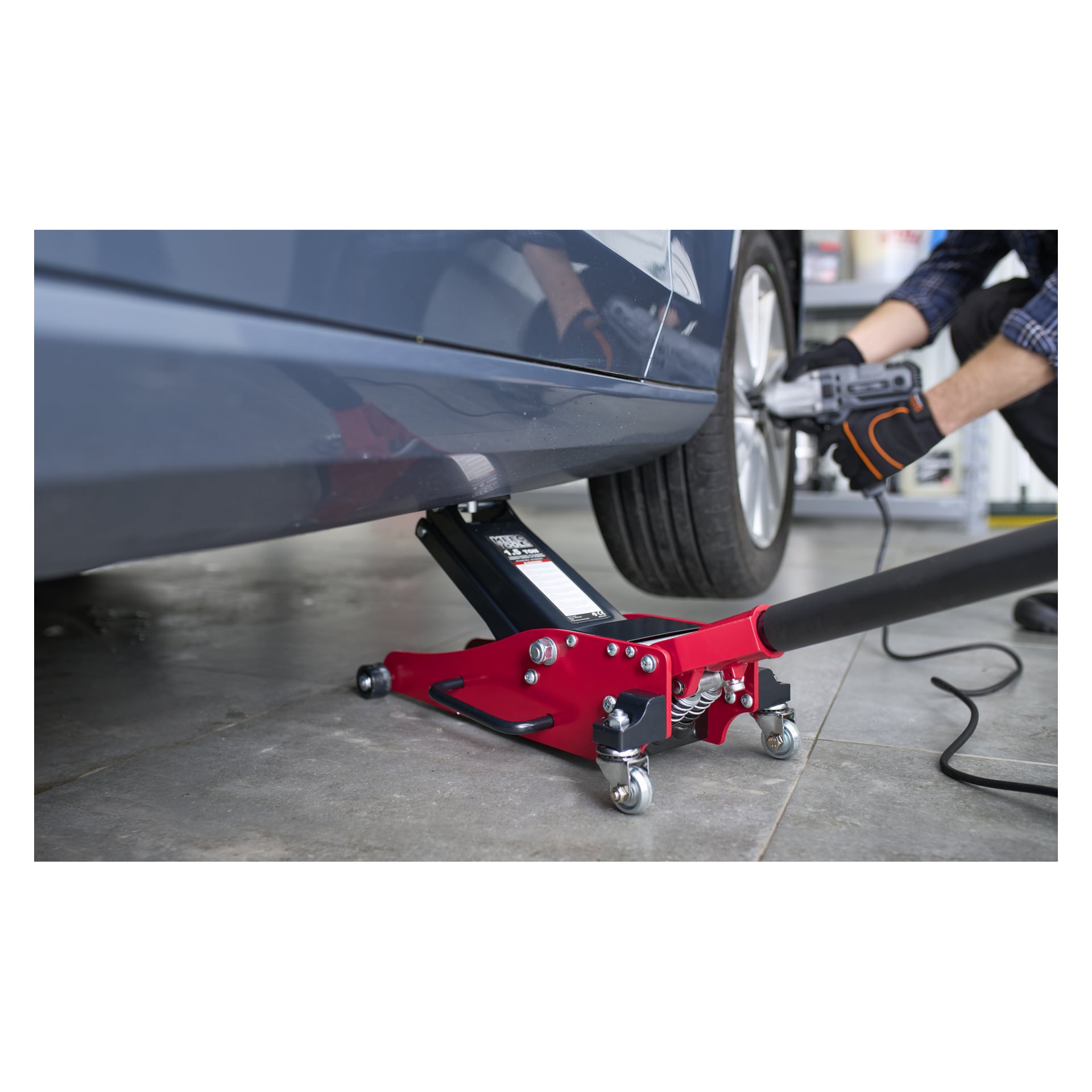 En bil är upphöjd på ett garagegolv med hjälp av en Meec Tools-domkraft med aluminiumchassi. En person med handske använder ett elverktyg för att arbeta med hjulmuttrarna på bilens framhjul.