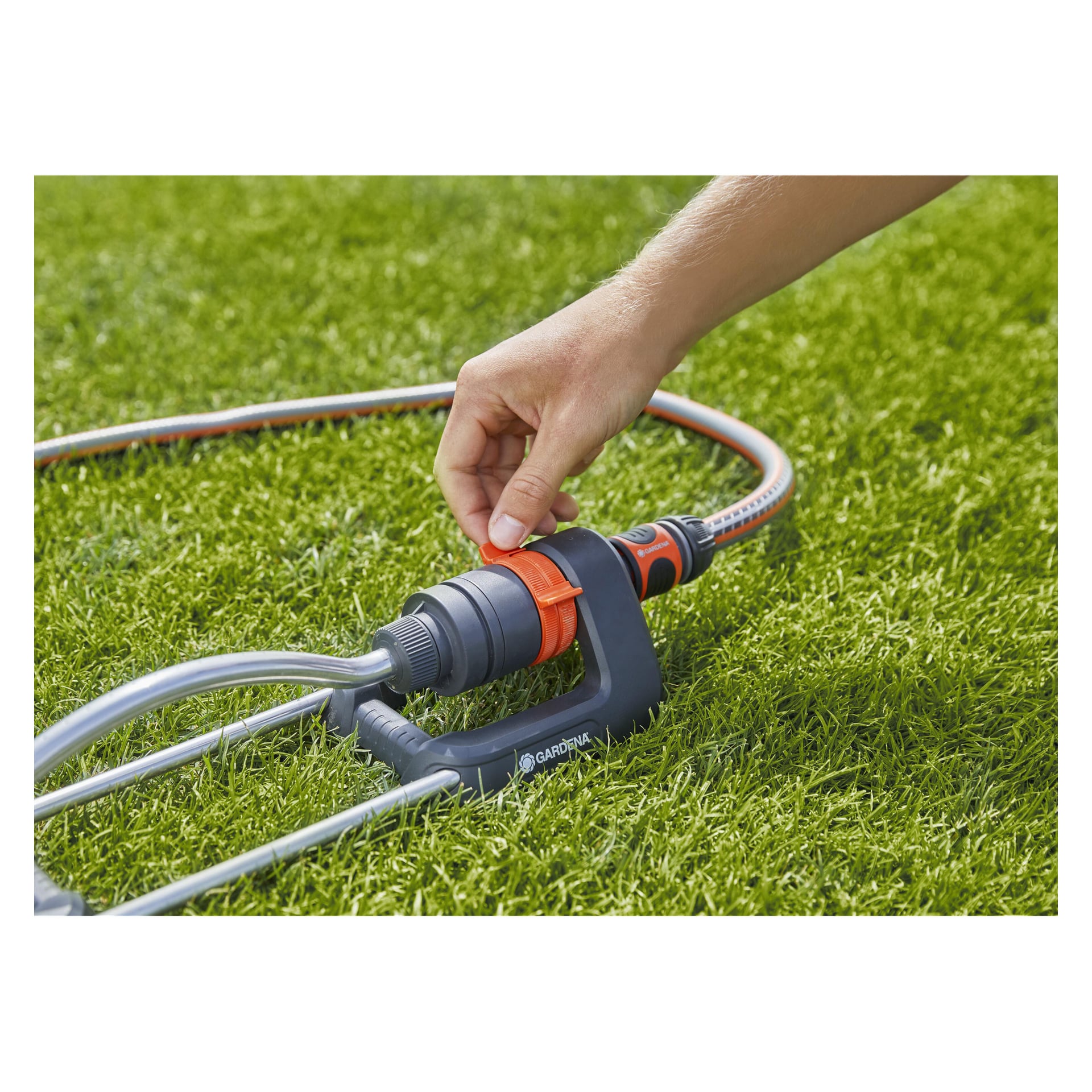 En hand vrider på den orangea ratten på en Gardena Aqua S, en svart och grå gräsmattesprinkler kopplad till en trädgårdsslang som vilar på grönt gräs.