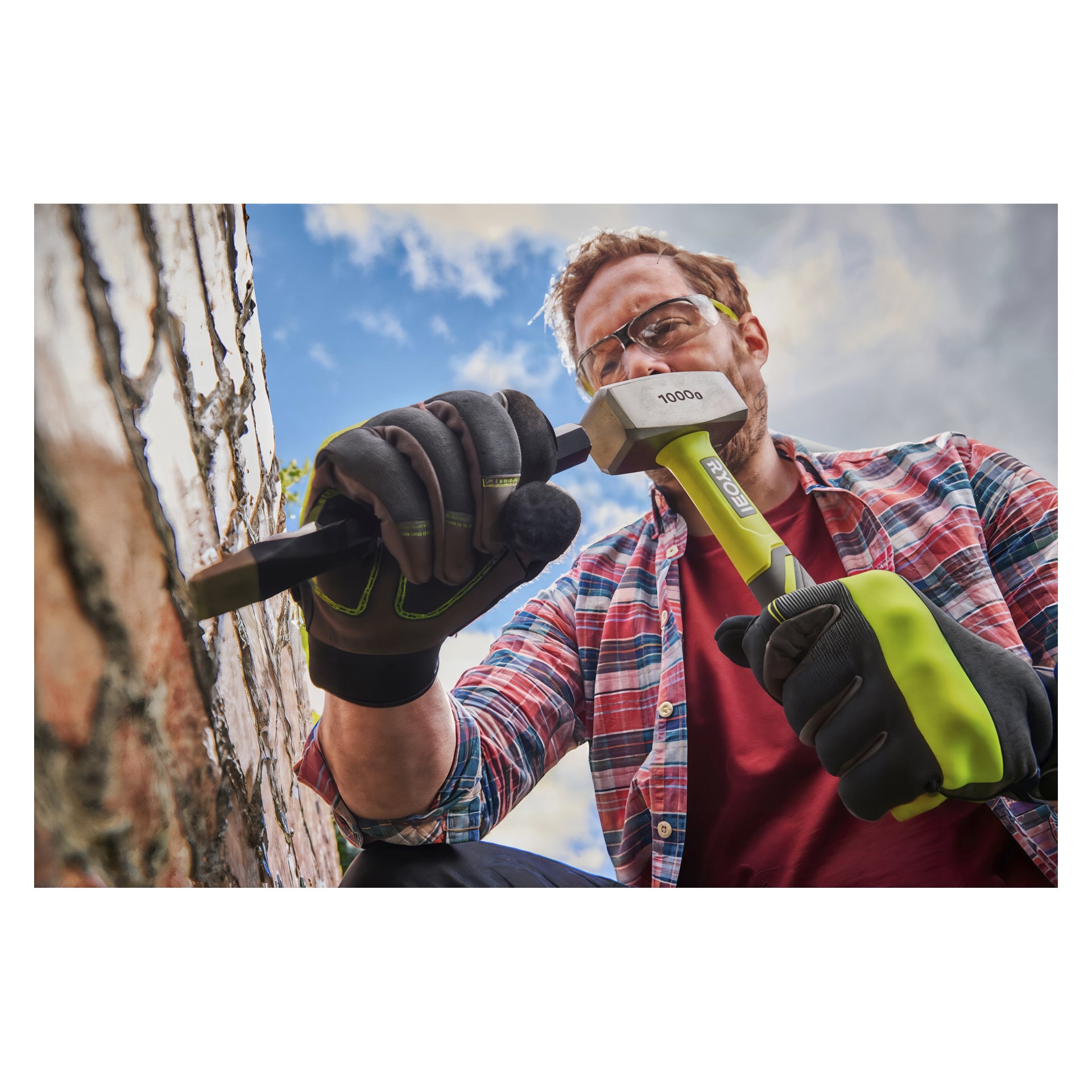 Med handskar och skyddsglasögon använder man Ryobi-mejselhammaren RHHCH1000 på en tegelvägg. Fotot är taget ur en låg vinkel med verktyg och säkerhetsutrustning tydligt synliga.