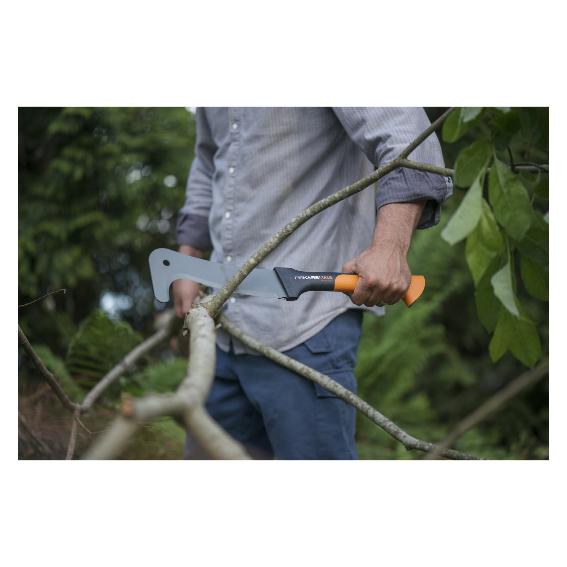 En person i grå skjorta och blå byxor är utomhus bland grön vegetation och klipper en trädgren med Fiskars Clearing Knife WoodXpert XA3 Medium.