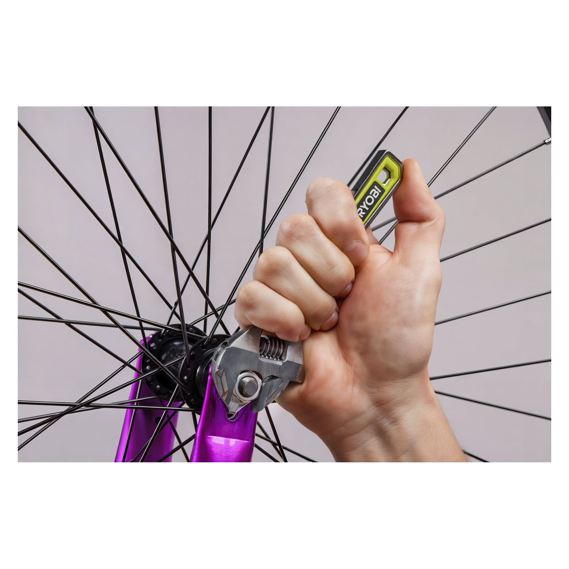 En hand använder en justerbar Ryobi-skiftnyckel på en bult fäst vid ett cykelhjul med svarta ekrar och en lila gaffel, placerad mot en diskret bakgrund.