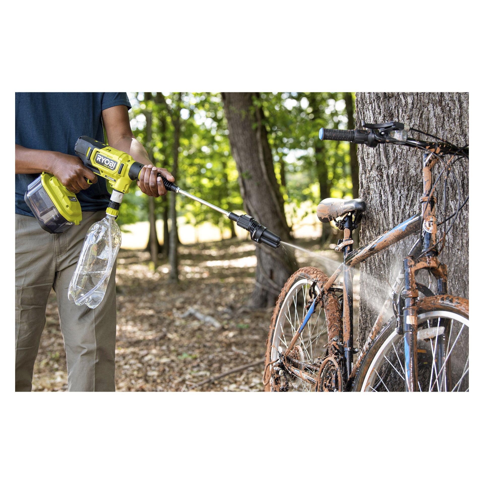 Någon använder en Ryobi One+ HP batteridriven medelhög högtryckstvätt (modell RY18PWX41A-0), utrustad med plastflaska, för att tvätta bort lera från en cykel lutad mot ett träd i en skogsklädd utomhusmiljö.