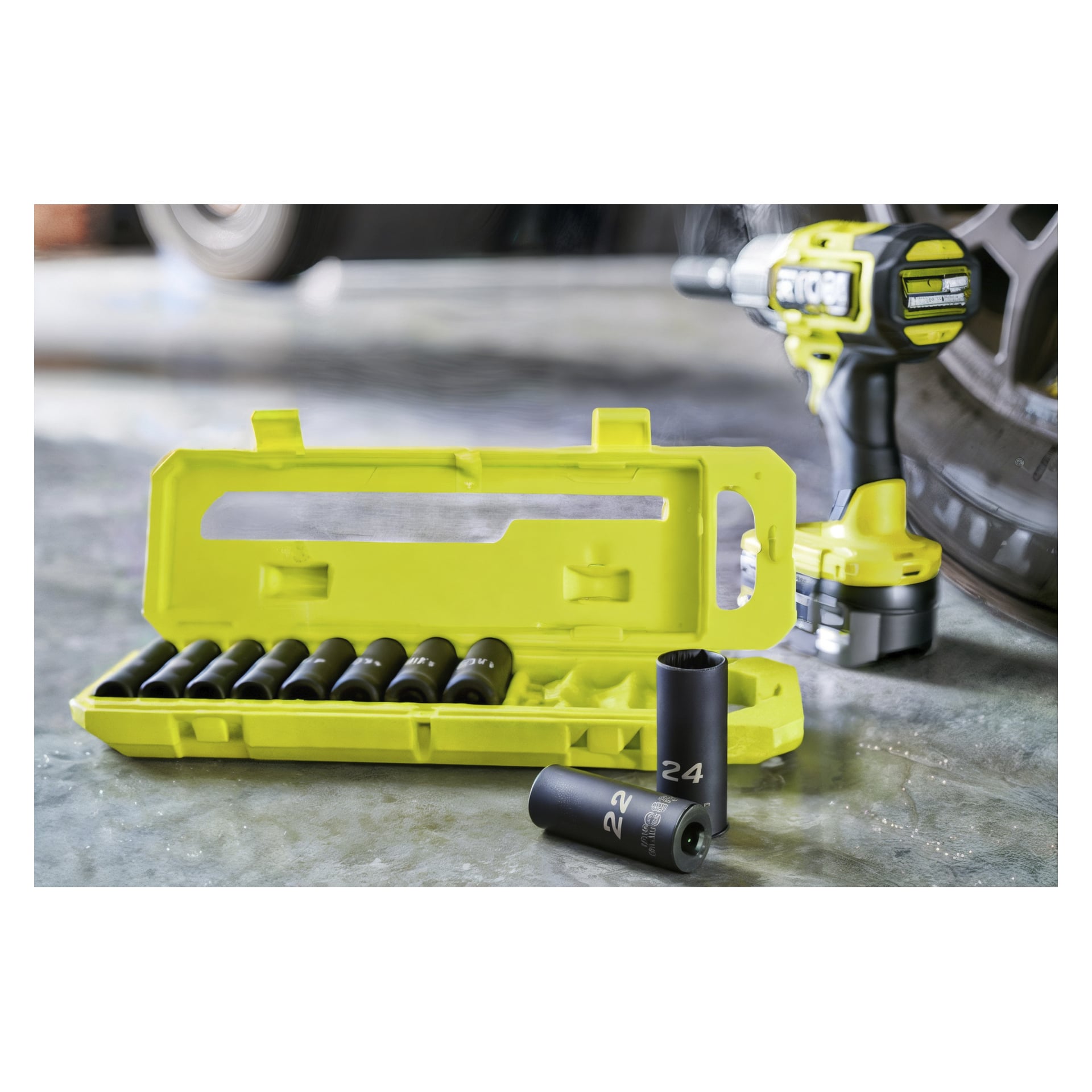Ett gult Ryobi-plastfodral innehåller ett 10-delat RHDIS10PC hylsset med svarta hylsnycklar, varav en står upprätt framtill. I bakgrunden ligger en slagnyckel nära ett bildäck på betonggolvet.