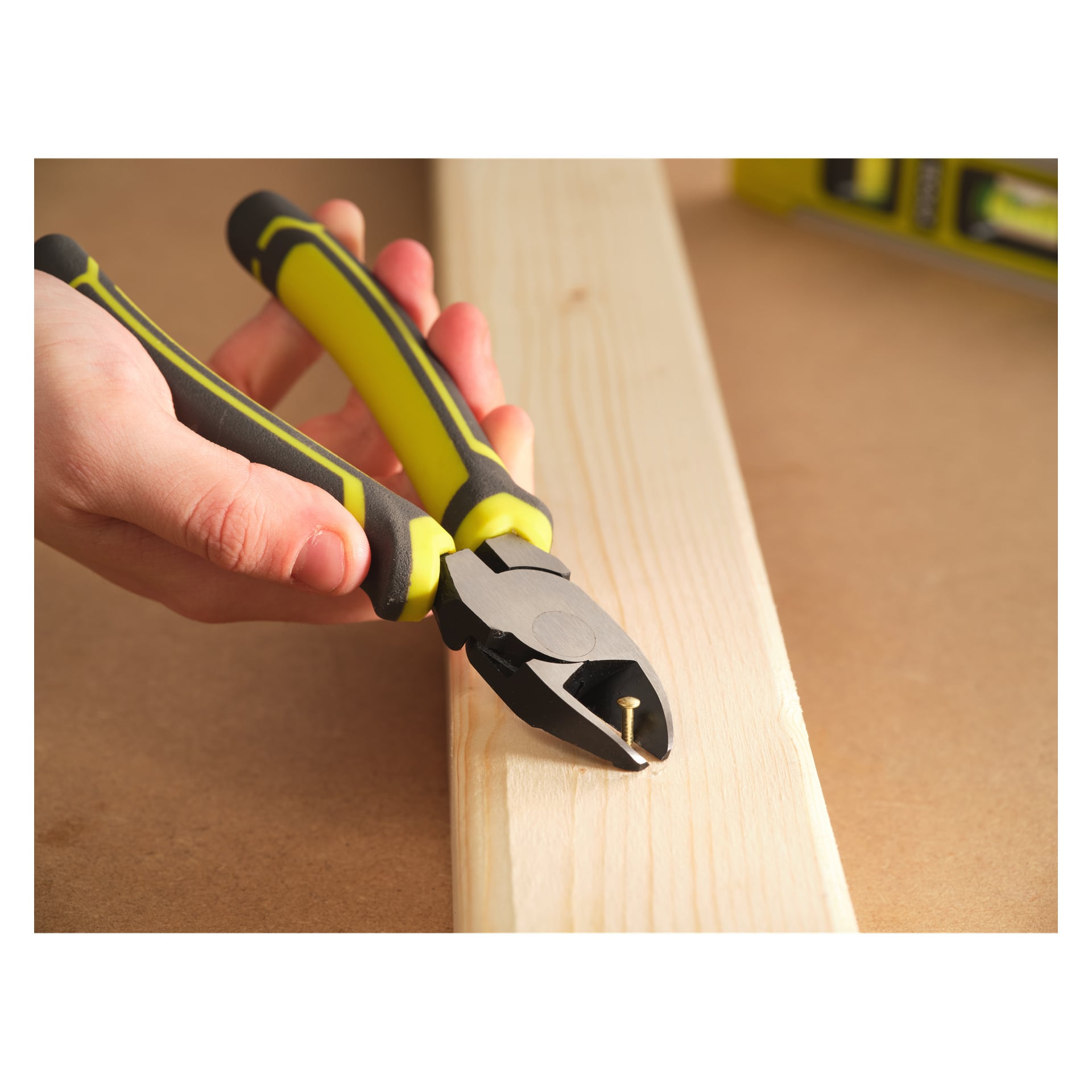 En hand använder Ryobi RHDC160 trådsax, med gult och svart färgschema, för att greppa en spik som delvis är inslagen i en ljus träskiva på ett arbetsbord.