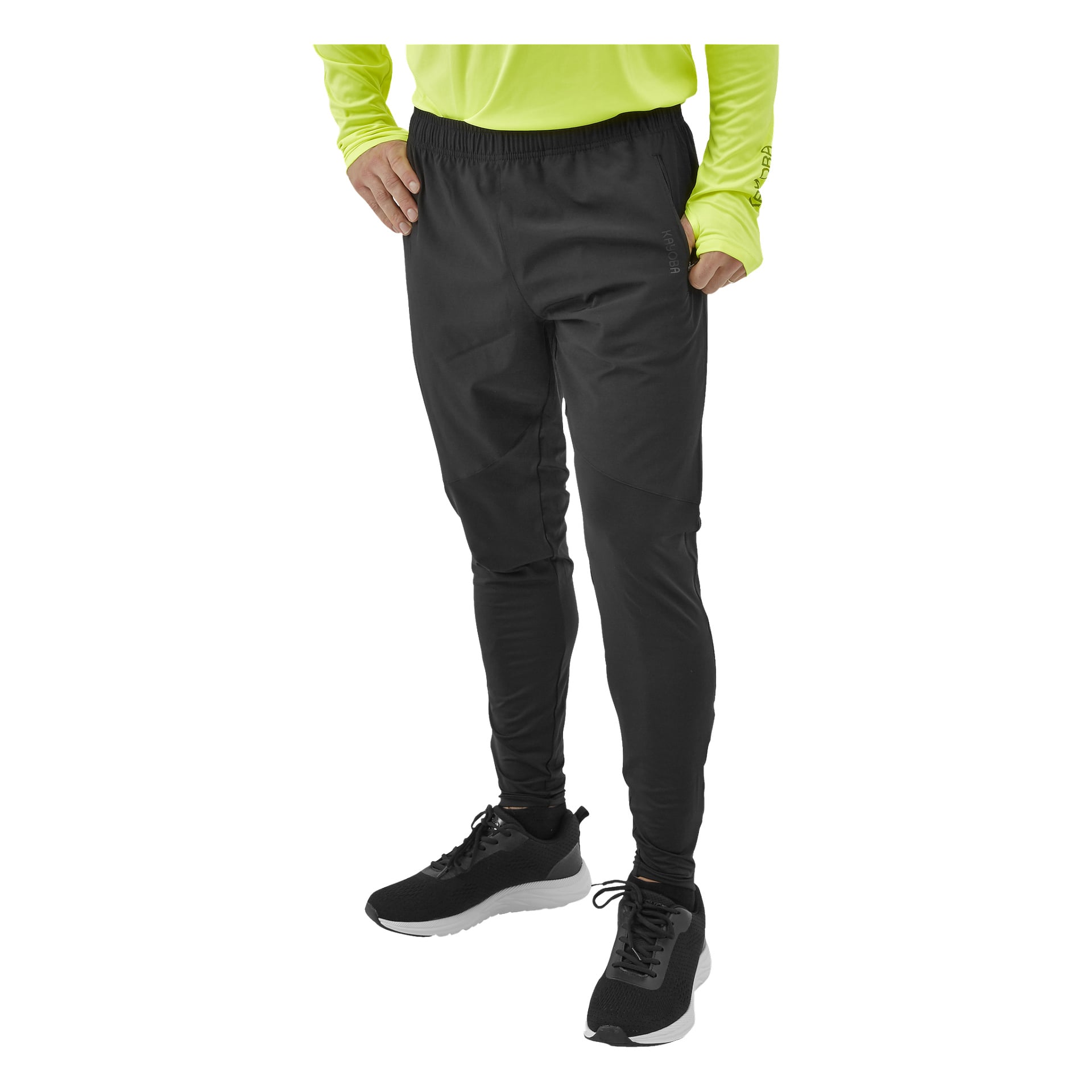 En person klädd i svarta Kayoba Men's Functional byxor Rio, en neongul långärmad skjorta och svarta sneakers med vita sulor står med ena handen i fickan.