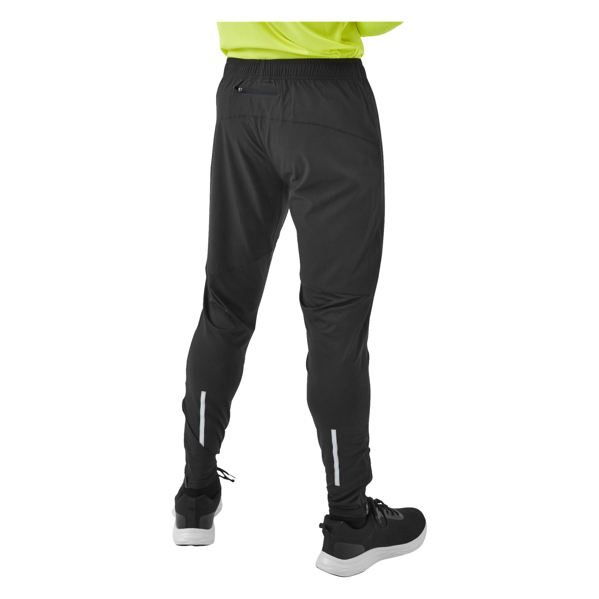 Sett bakifrån bär man Kayoba Men's funktionella byxor Rio, som har en bakficka med dragkedja och reflekterande remsor vid anklarna. De bär också svartvita sneakers, med en del av en neongrön tröja synlig upptill.