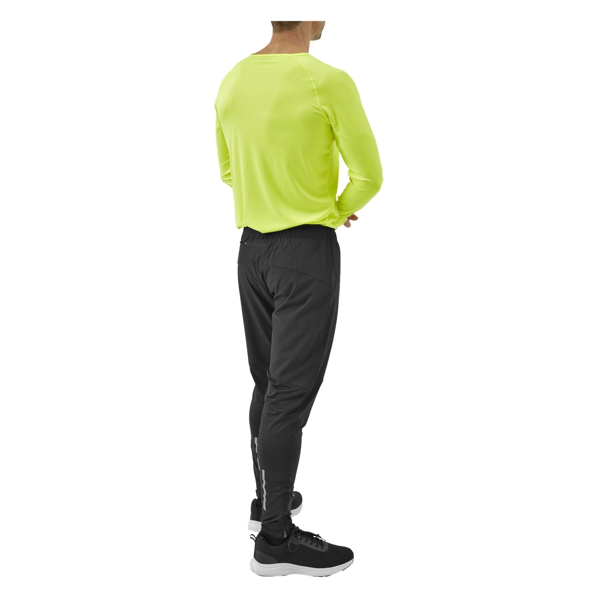 En person står med ryggen mot kameran, klädd i en livfull gulgrön långärmad skjorta, Kayoba Men's funktionella byxor Rio och svarta sneakers med vita sulor.