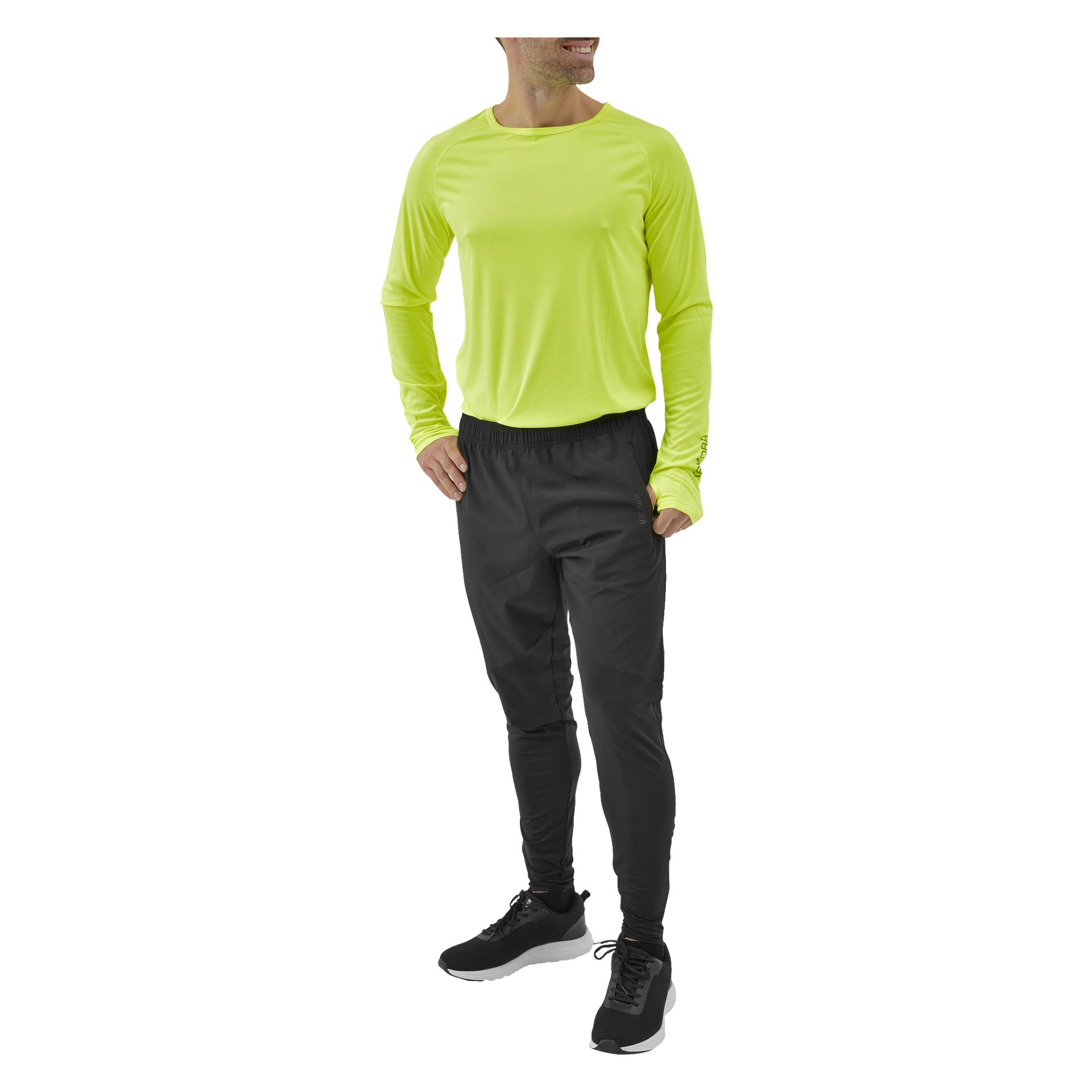 En person med händerna i fickorna är klädd i en neongul långärmad skjorta, svarta Kayoba herrbyxor i Rio och svarta sneakers. De ler och tittar åt sidan.