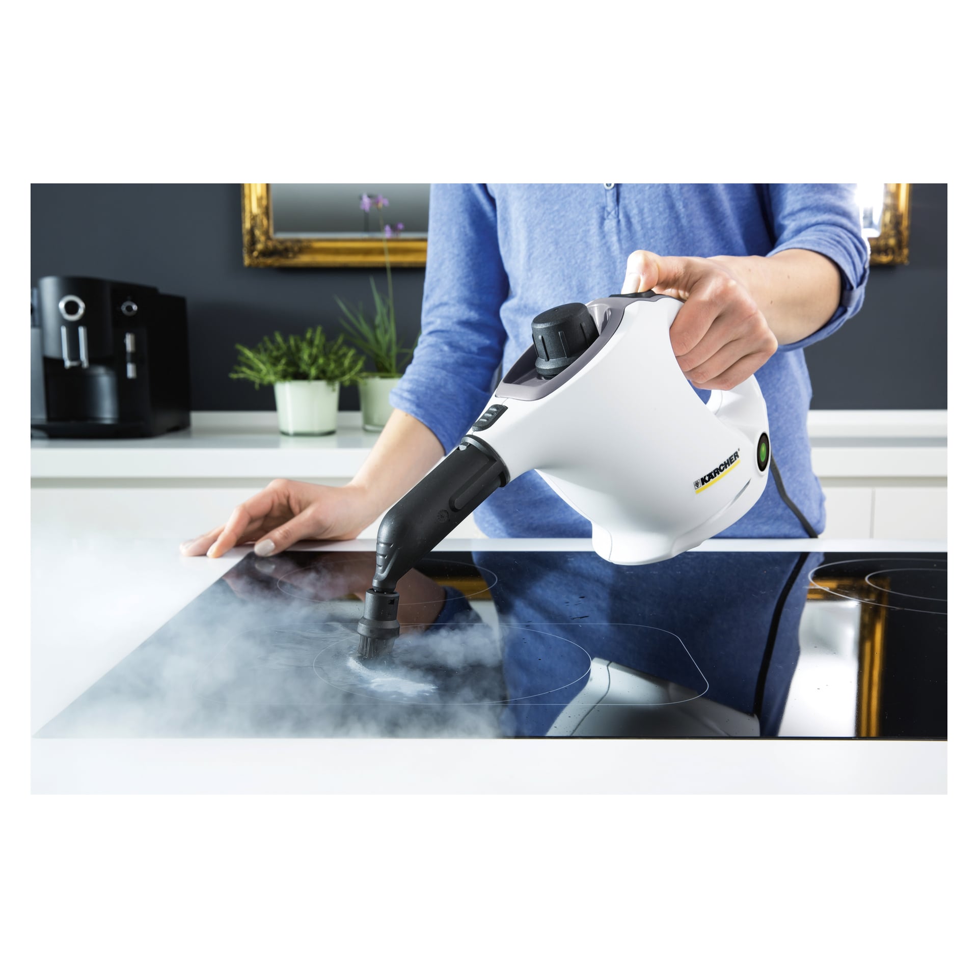 Någon rengör en spishäll i ett modernt kök med Kärcher Steam Cleaner SC 1 7-delat set, med ånga tydligt synlig från apparaten. I bakgrunden finns en växt, några köksapparater och en spegel.