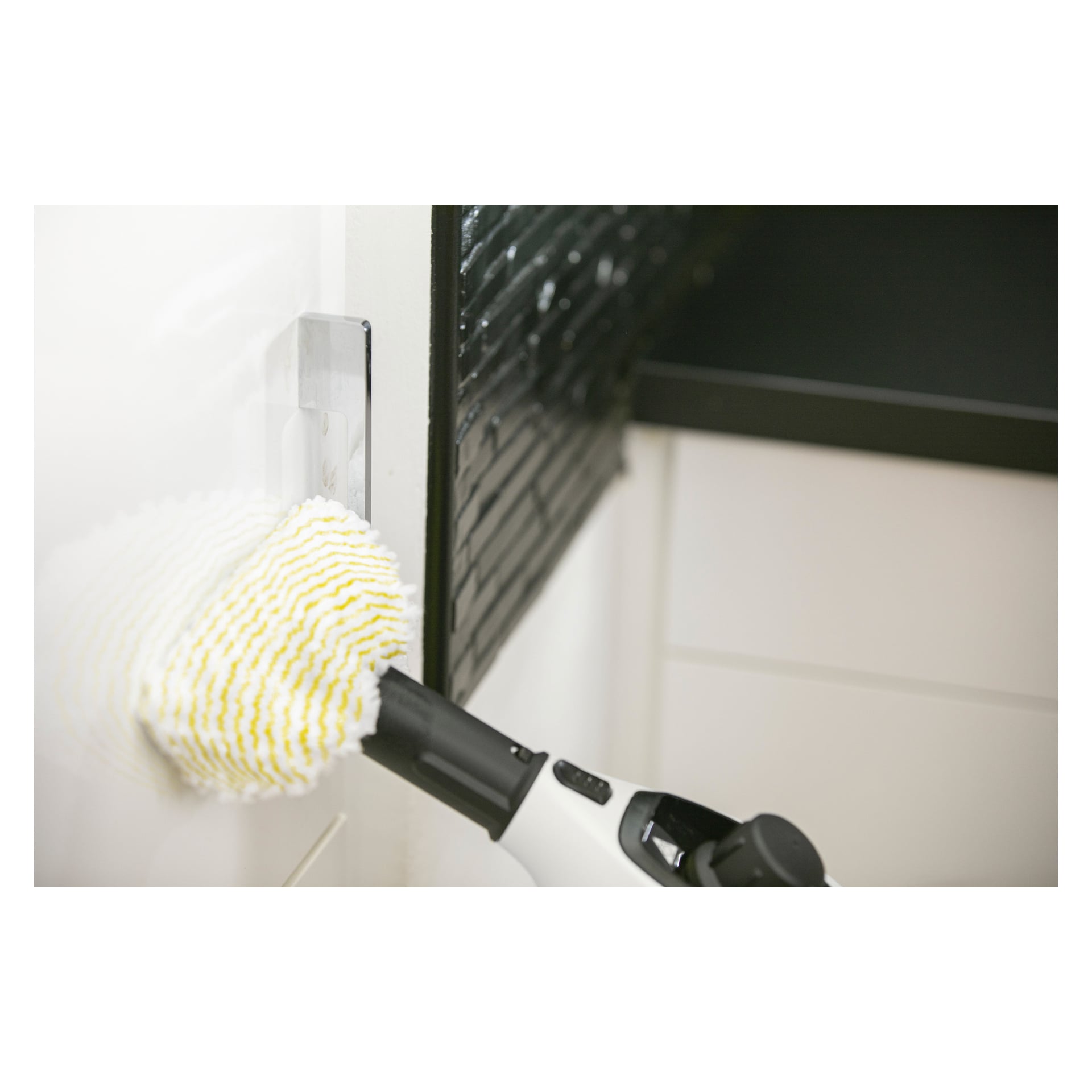 Någon rengör en vit kaklad vägg med Kärcher Steam Cleaner SC 1 7-delat set, med ett vitt och gult rengöringsverktyg, bredvid en svart stänkskydd och vita skåp i ett kök eller badrum.