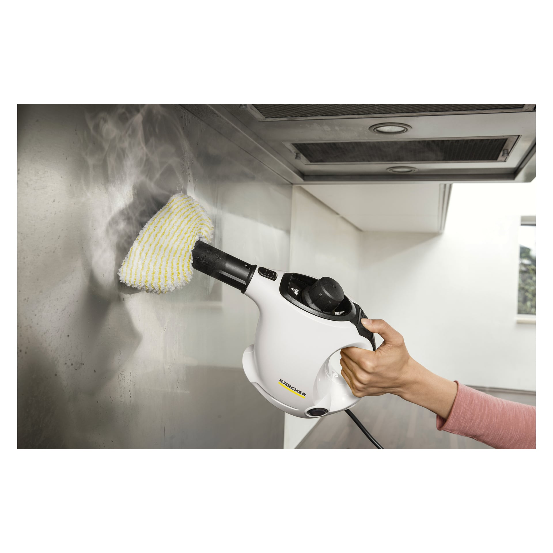 Man rengör en köksyta i rostfritt stål med Kärcher Steam Cleaner SC 1, med hjälp av en tygbit. Ånga är synlig från apparaten i en modern köksmiljö.
