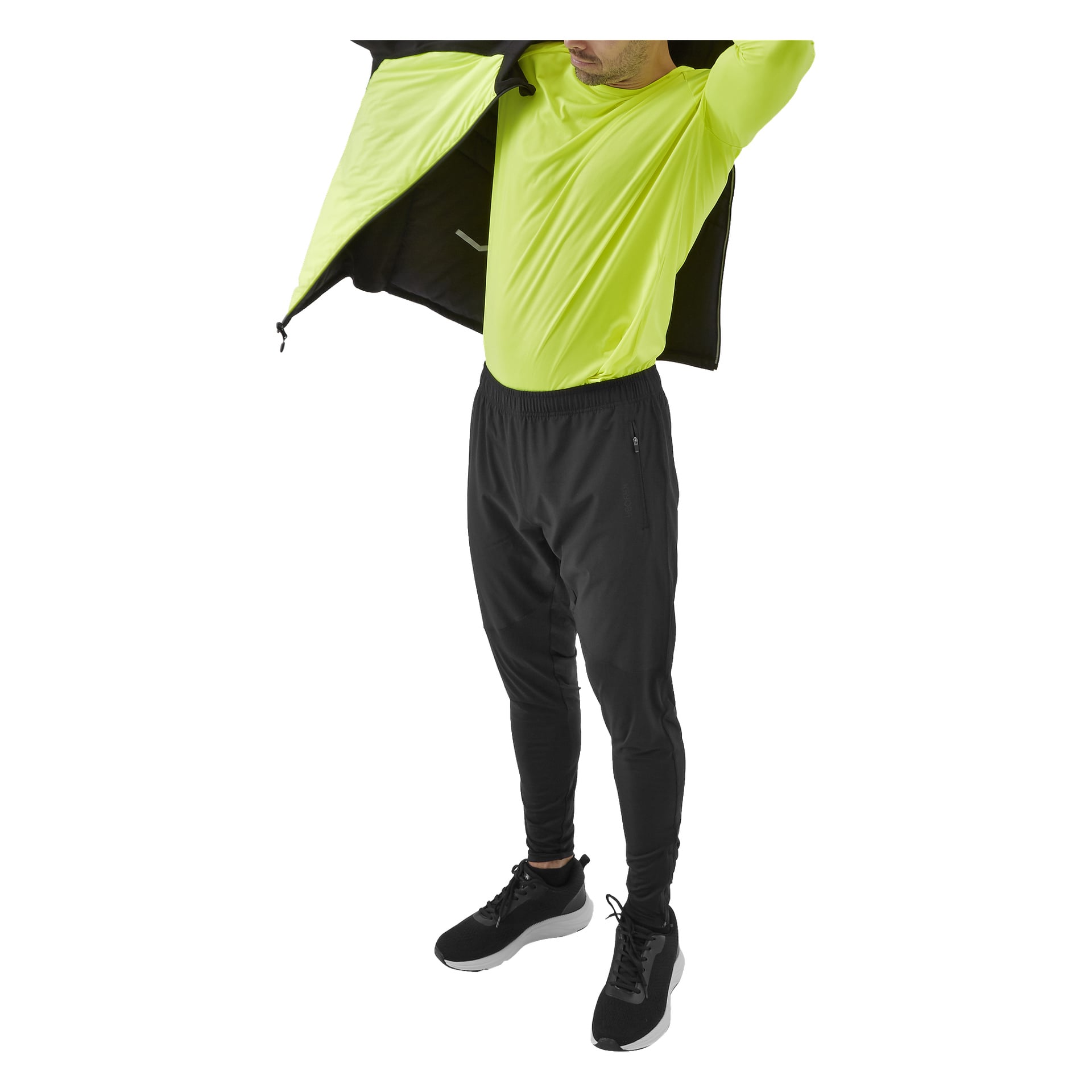 En person drar Kayoba Reversible Innsbruck löparjacka för män över axlarna. De bär en neongul långärmad skjorta, svarta byxor och svarta sneakers. Den övre delen av deras huvud är beskuren ur bilden.