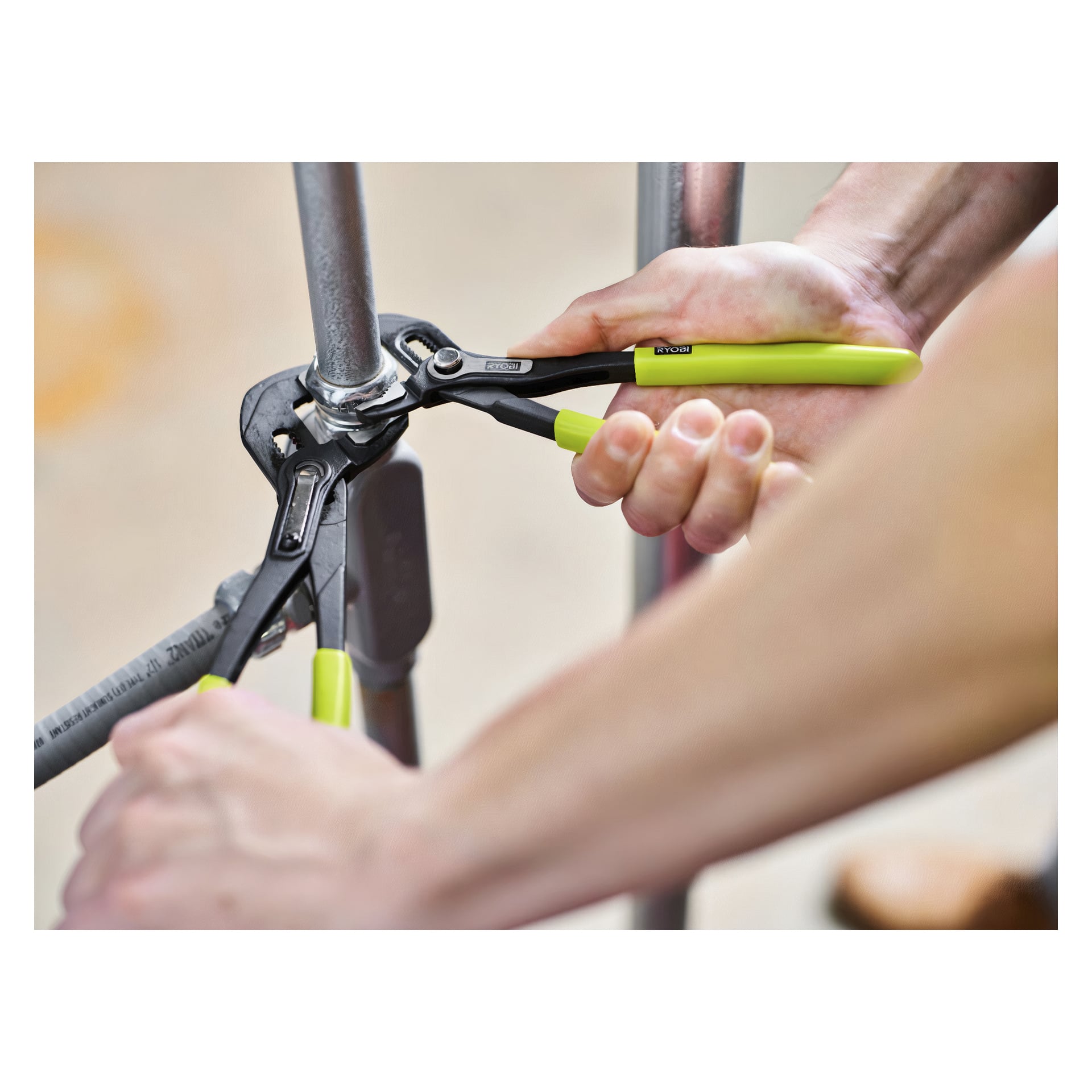 Ett par händer greppar grönskaftade Ryobi Polygrip-tånger och arbetar på en metallrörsfitting. Bakgrunden är suddig, vilket betonar aktiviteten och verktygen.