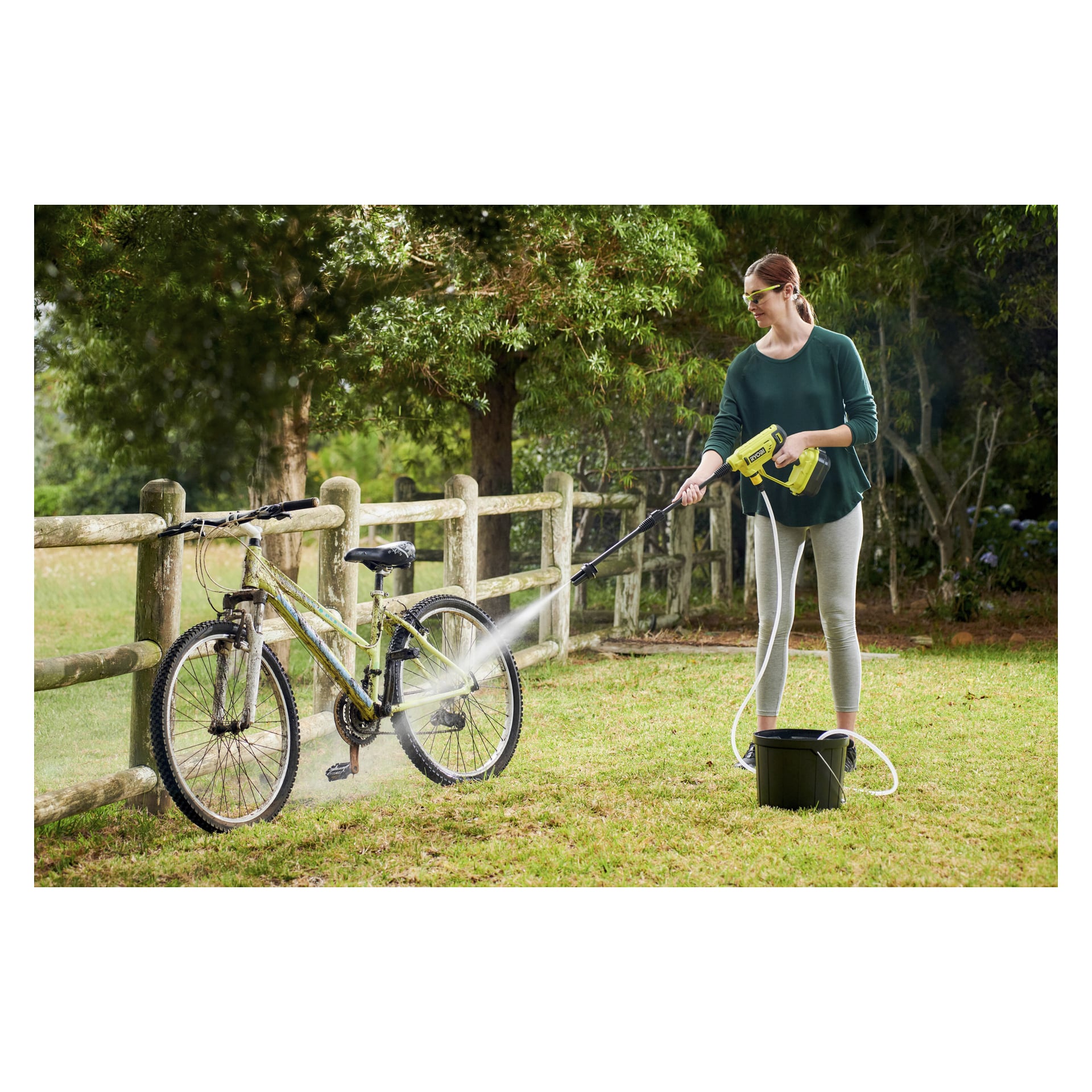 Iklädd en grön skjorta och grå byxor använder man Ryobi One+ batteridriven medelhög högtryckstvätt (modell RY18PW22A-125) för att tvätta en grön cykel på gräsbevuxen mark, nära ett trästaket och träd. En svart hink står placerad i närheten på gräset.