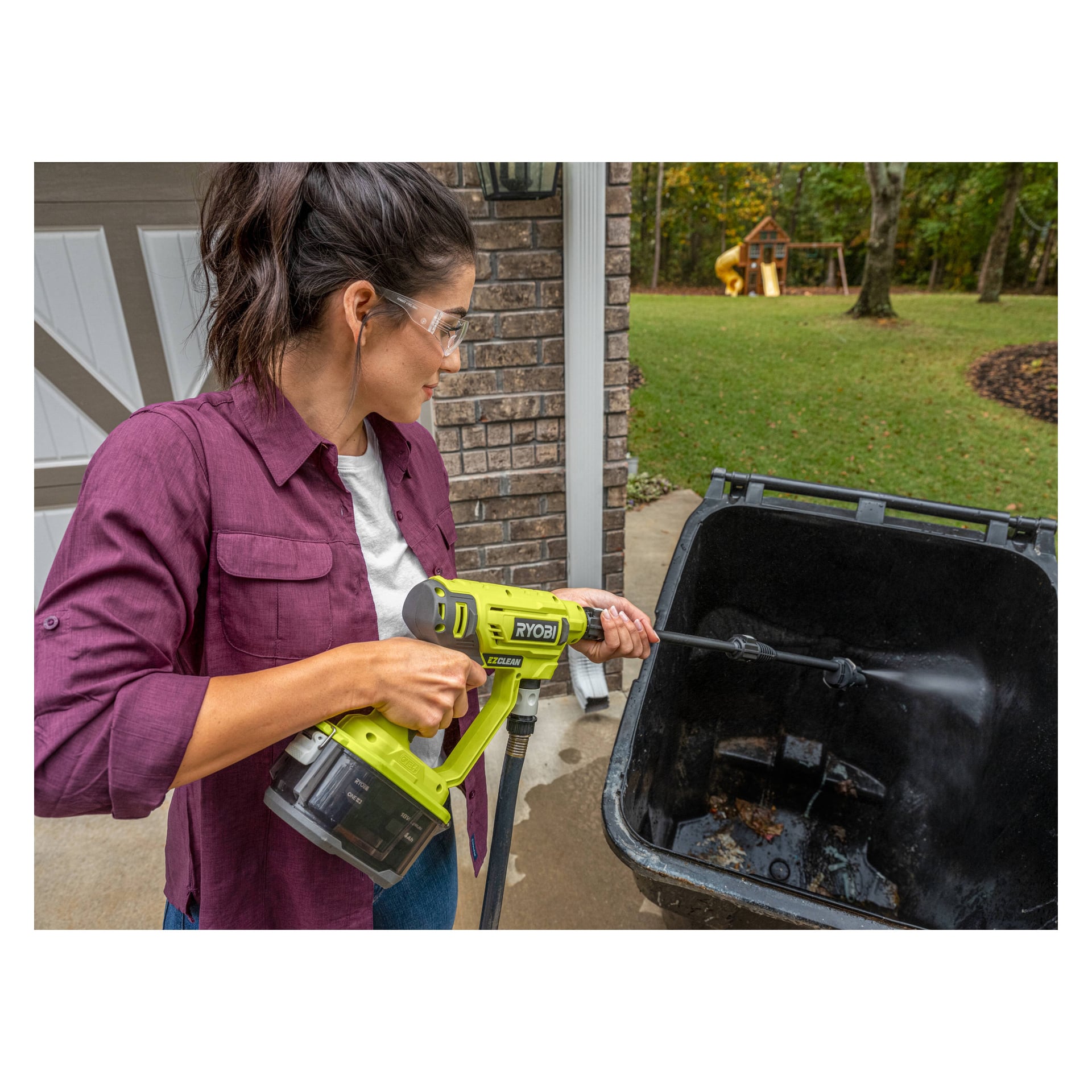 En kvinna med skyddsglasögon städar insidan av en stor utomhussoptunna med en Ryobi One+ batteridriven medelhögtryckstvätt (modell RY18PW22A-125) bredvid ett tegelhus. I bakgrunden finns en lekplats på en gräsbevuxen gräsmatta.