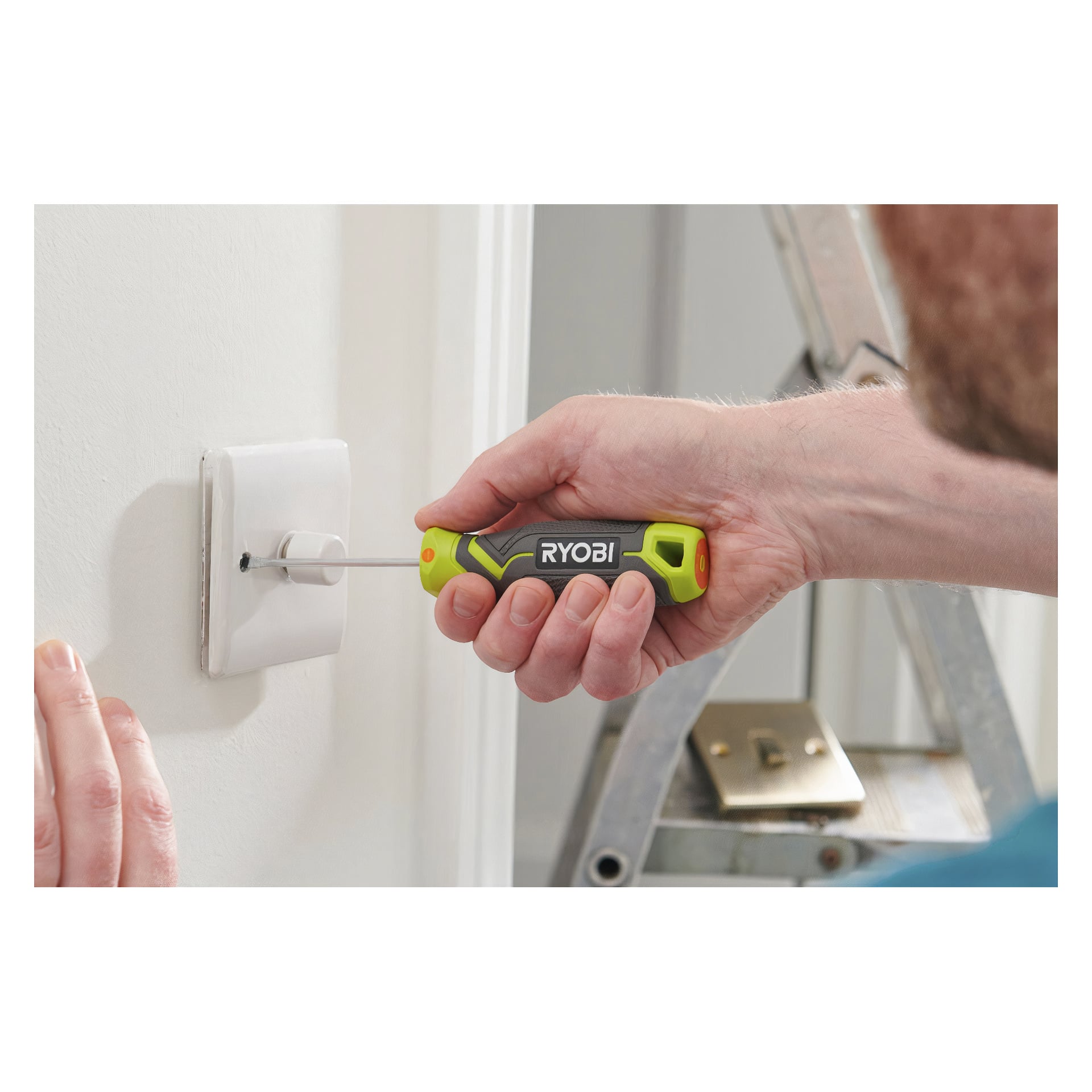 Någon skruvar loss en skruv från ett eluttagslock på väggen med hjälp av ett Ryobi RHSDS6PC sexdelat skruvmejselset, med en del av en metallstege synlig i bakgrunden.