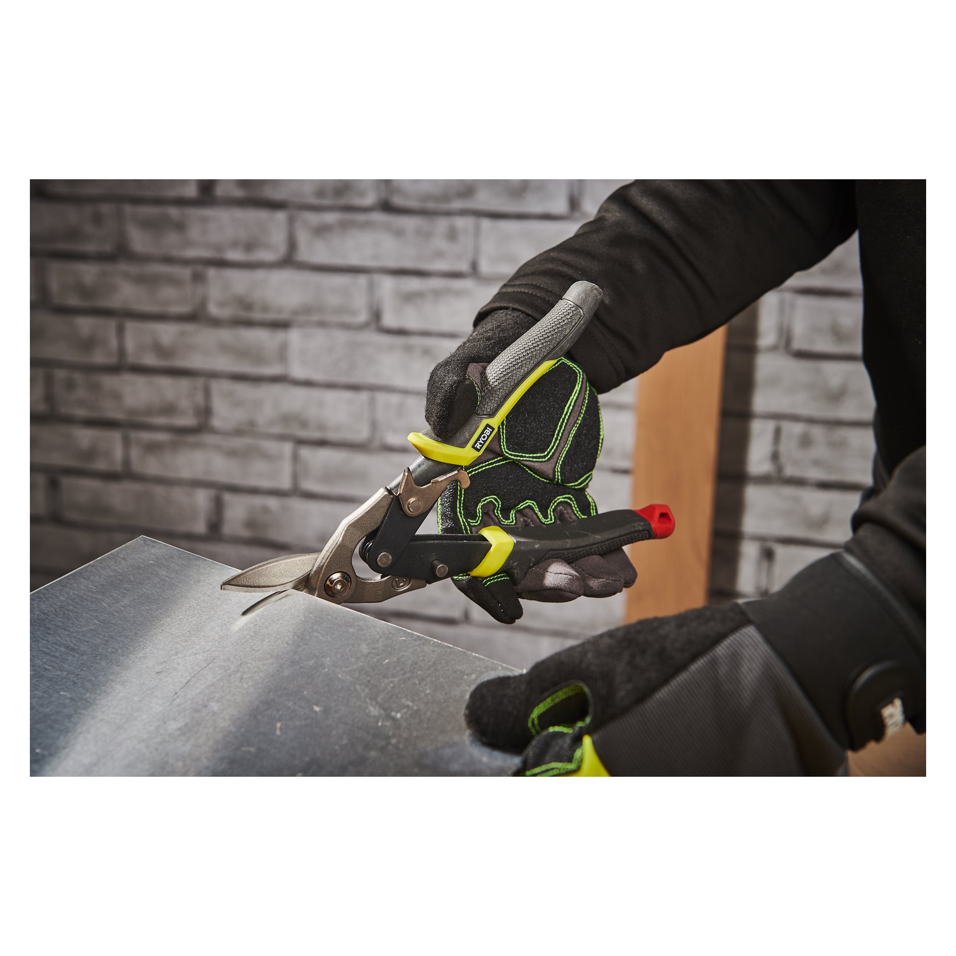 En person med skyddshandskar använder Ryobi Tin Saxs RHASL för att skära igenom en metallplåt, med en grå tegelvägg och träplanka synliga i bakgrunden.