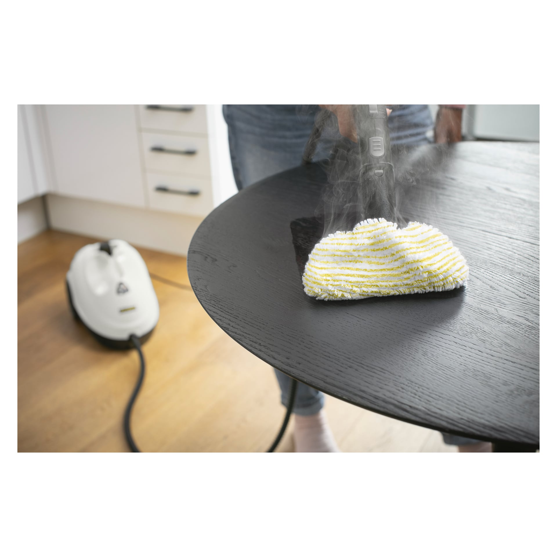 Någon rengör ett mörkt träbord med en Kärcher Steam cleaner SC 2 EasyFix med en randig trasa. Huvudenheten står på golvet, ansluten med en sladd.