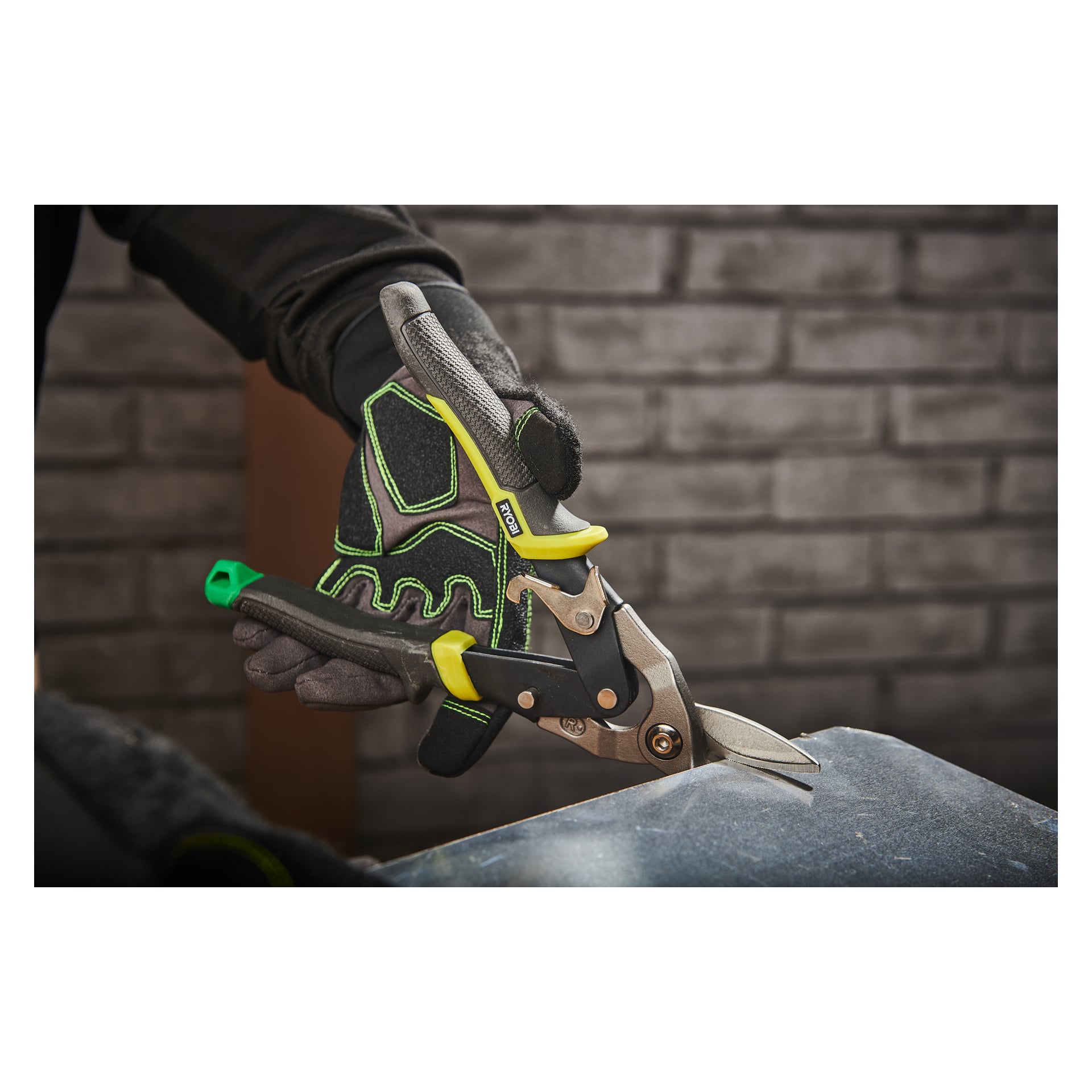 En person som bär handskar använder Ryobi Metal Saxs RHASR för att skära en plåt. Verktyget har gula och gröna handtag, med en grå tegelvägg i bakgrunden.