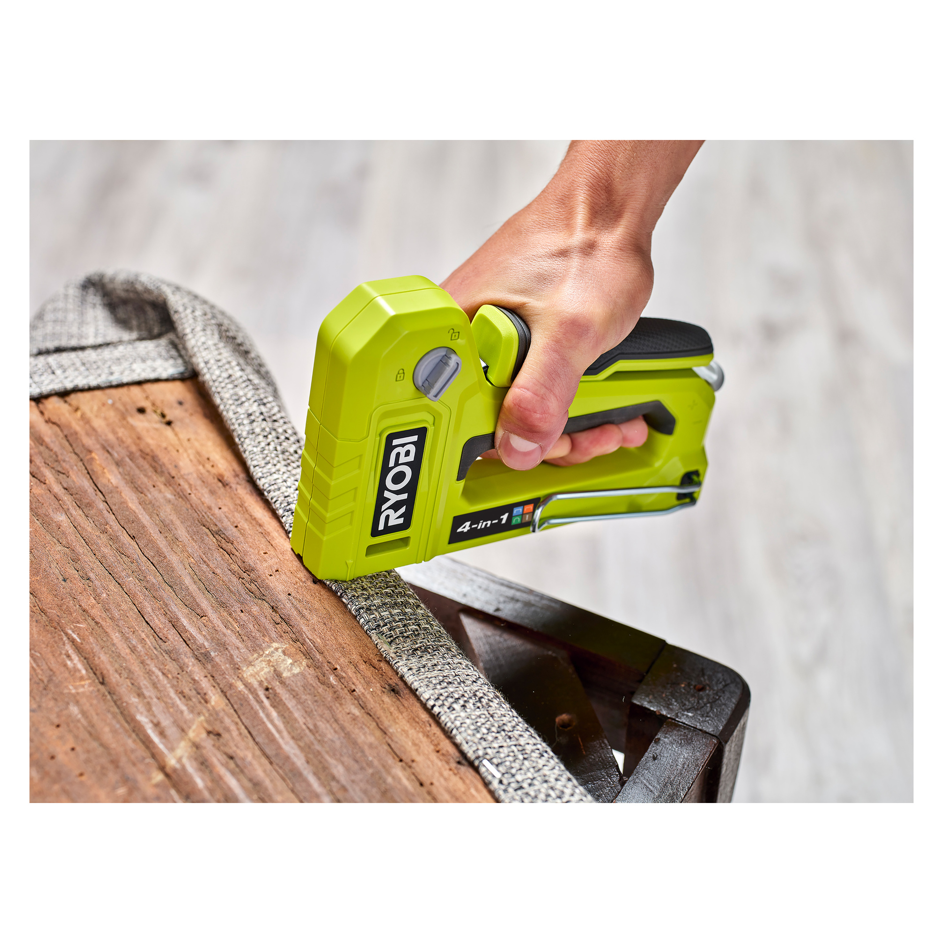En hand griper tag i en limegrön Ryobi 4-i-1 häftpistol RHFST4IN1 och trycker den längs kanten av det grå texturerade tyget som är lindat runt en trämöbelram.