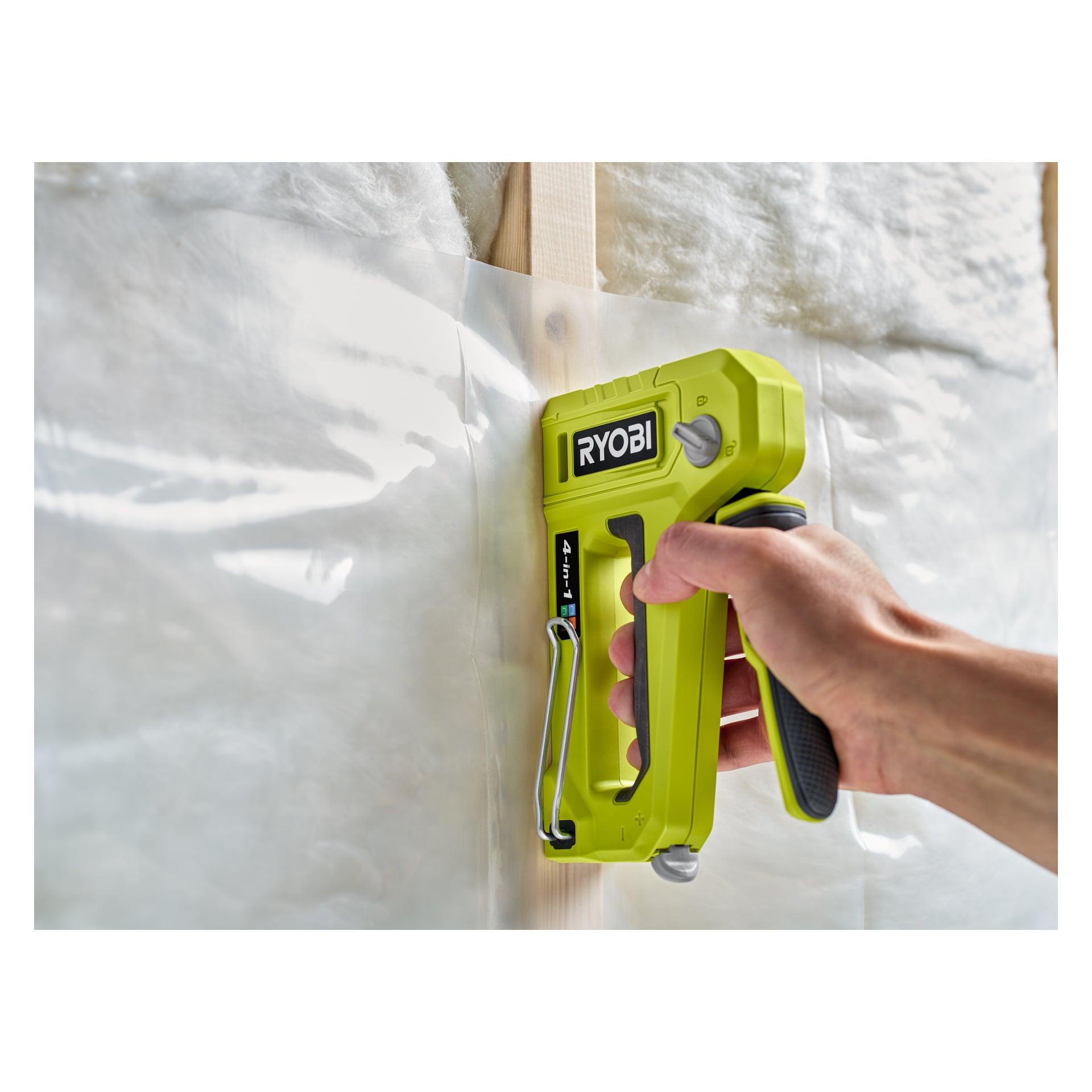 En hand håller i en Ryobi 4-i-1 häftpistol (modell RHFST4IN1), och fäster en plastångspärr på en träregla, med glasfiberisolering synlig i bakgrunden.