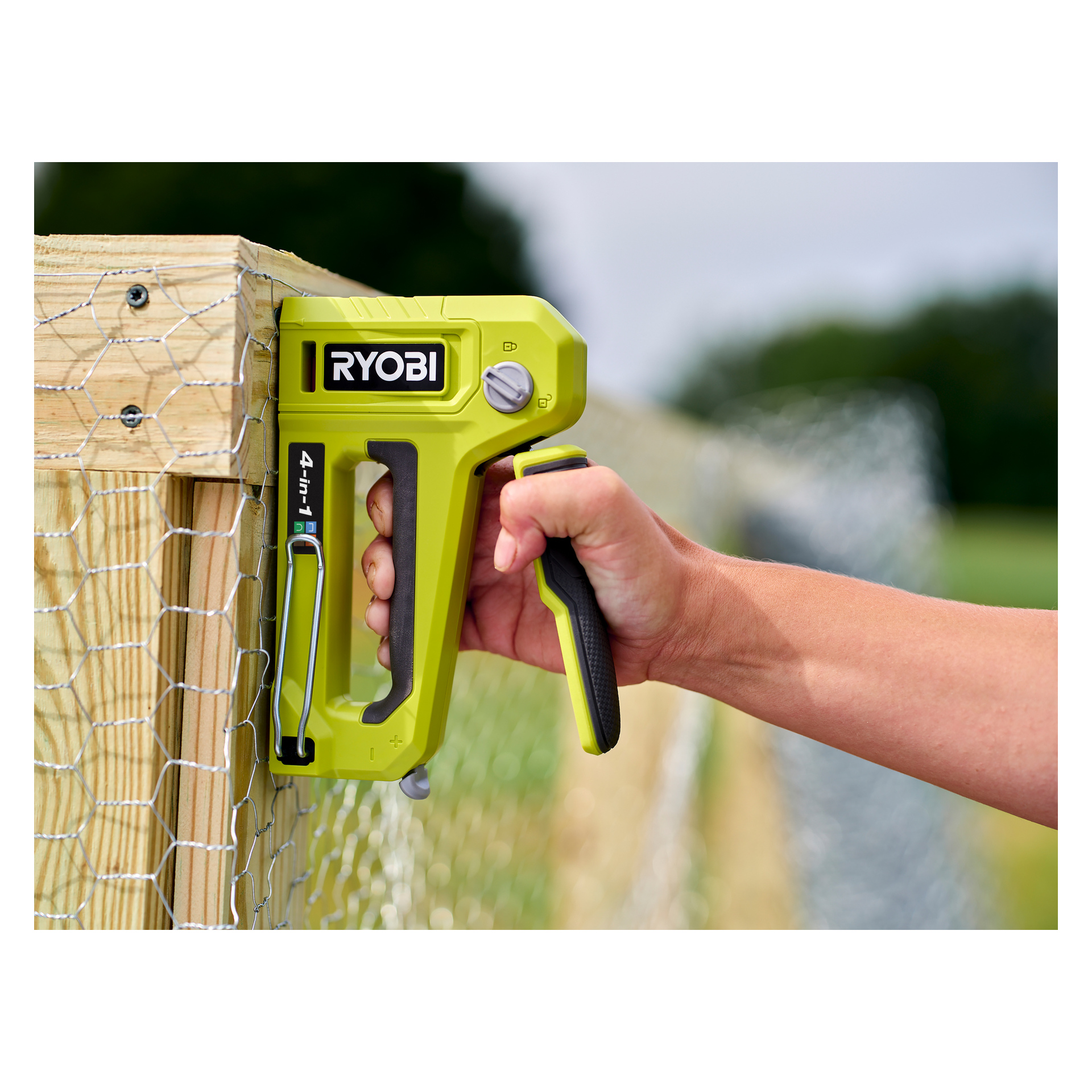 En person fäster trådnät på en träram utomhus med hjälp av Ryobi 4-i-1 häftpistol RHFST4IN1, med gräs och träd mjukt ur fokus i bakgrunden.
