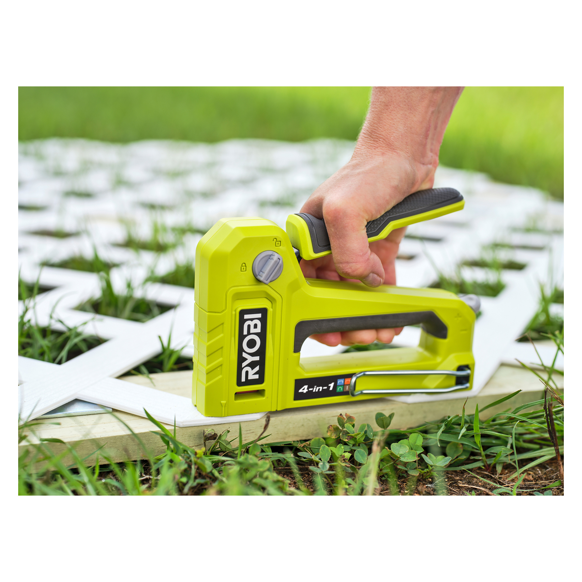 En hand greppar en limegrön Ryobi 4-i-1 häftpistol (modell RHFST4IN1) och trycker den mot en vit plastgallerpanel placerad på en träplanka utomhus på gräset.