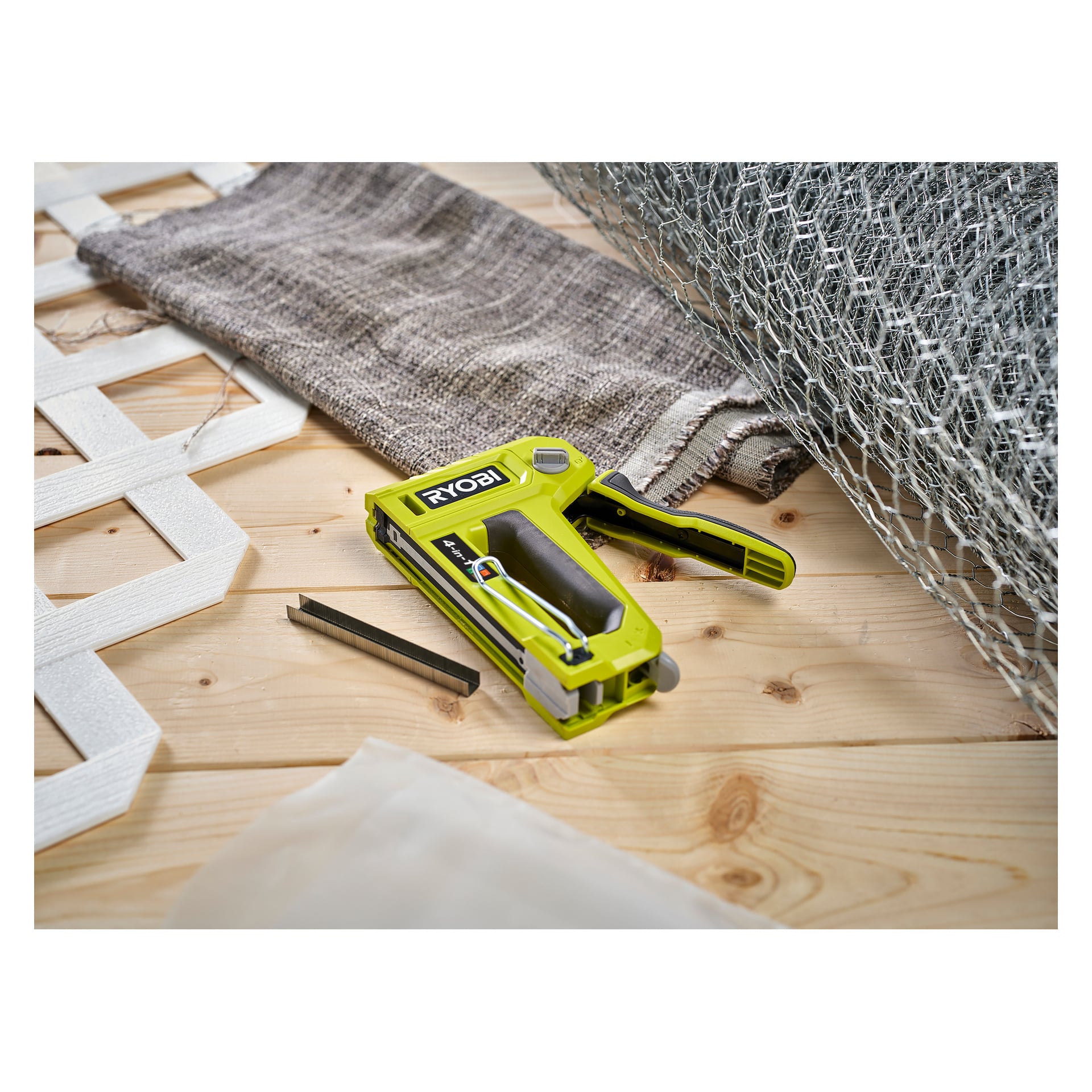 En livfull grön Ryobi 4-i-1 häftpistol, modell RHFST4IN1, ligger på en träyta med tyg, metallnät, en rektangulär metallremsa och ett vitt gallerstycke placerat i närheten.