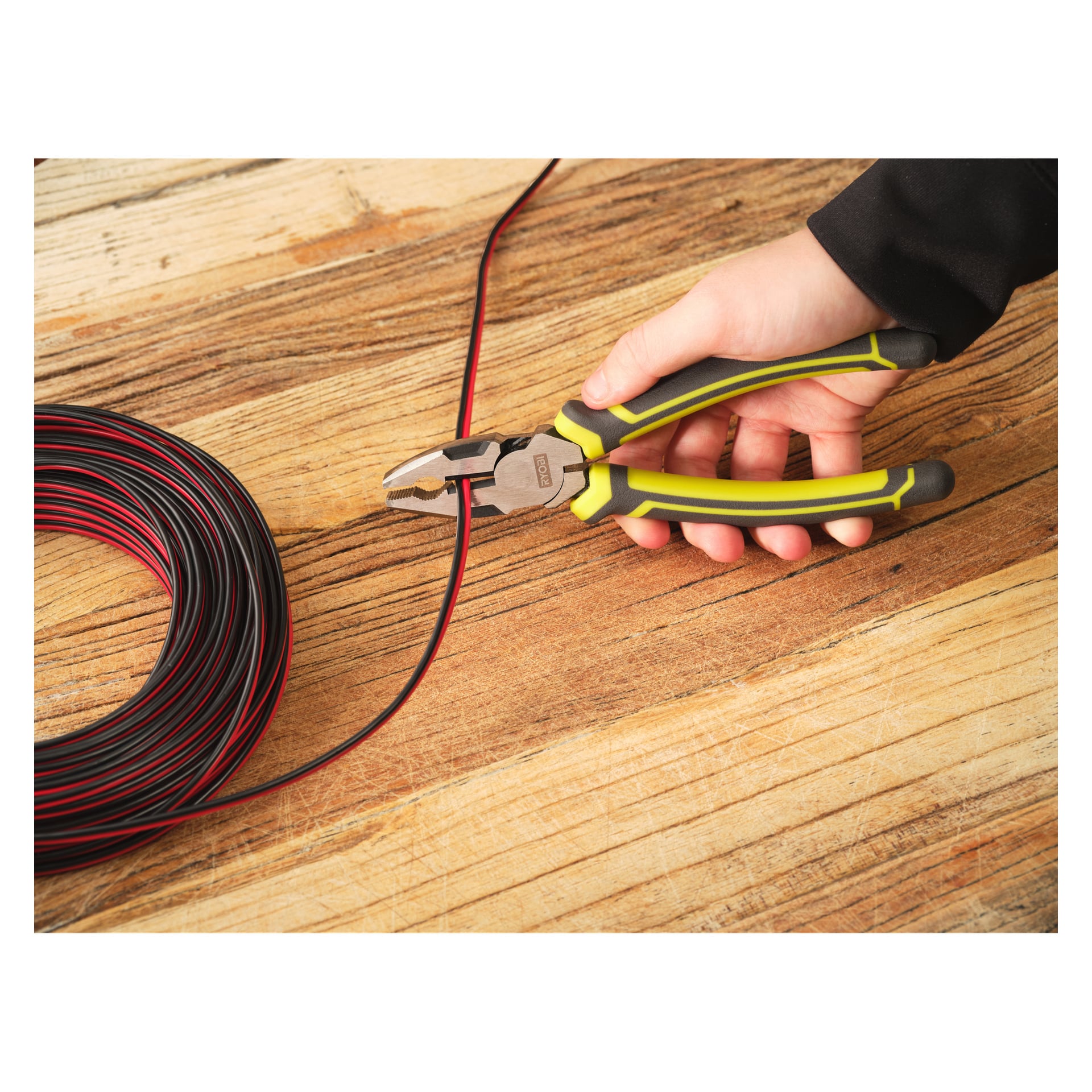 En hand använder Ryobi Combination Pliers RHCP180 för att klippa en röd och svart elektrisk tråd, medan en spole av samma tråd ligger på ett träbord.