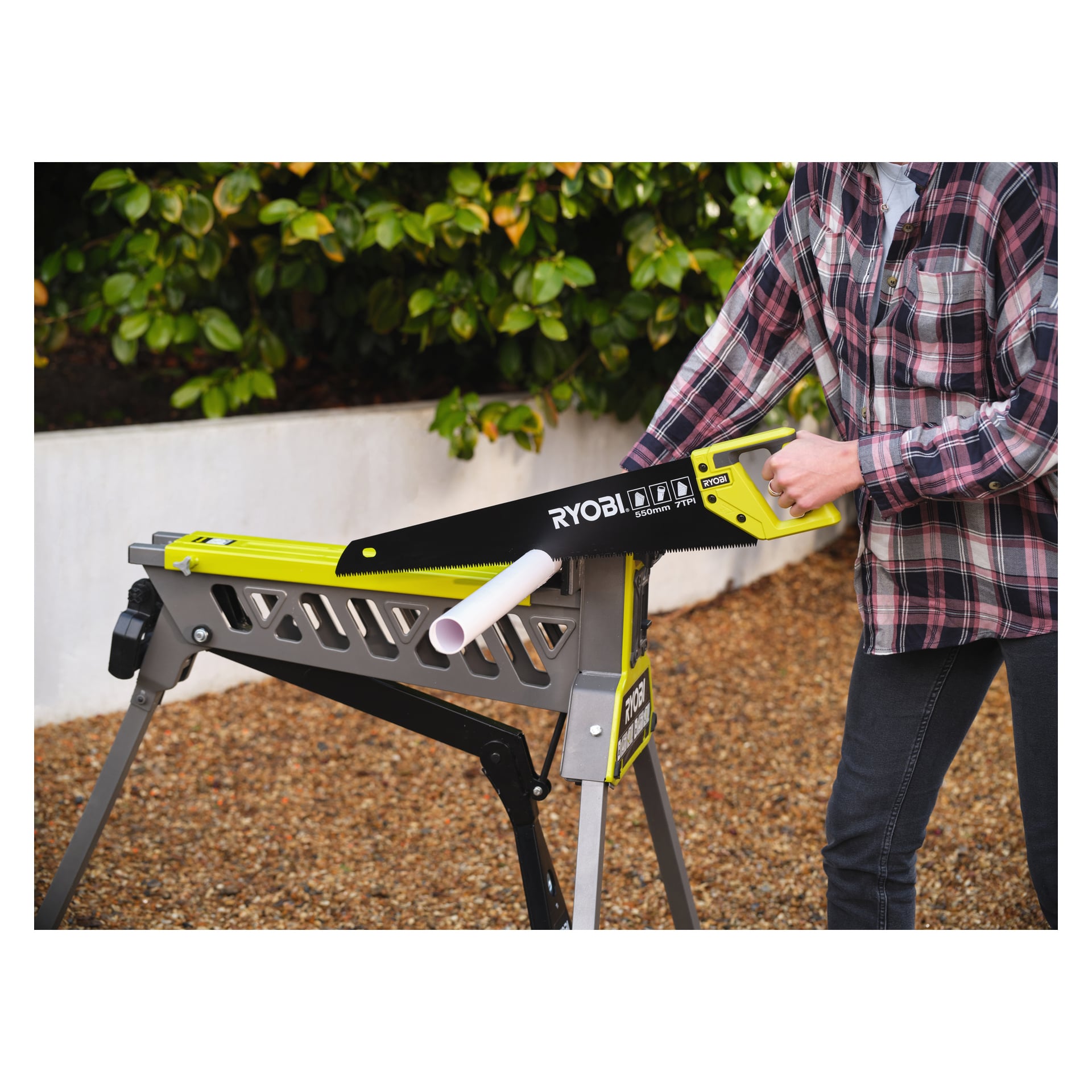En person i rutig skjorta och jeans använder Ryobi Handsaw RHCHS-550 för att skära ett vitt plaströr som vilar på en metallsåg utanför, med buskar och grus i bakgrunden.