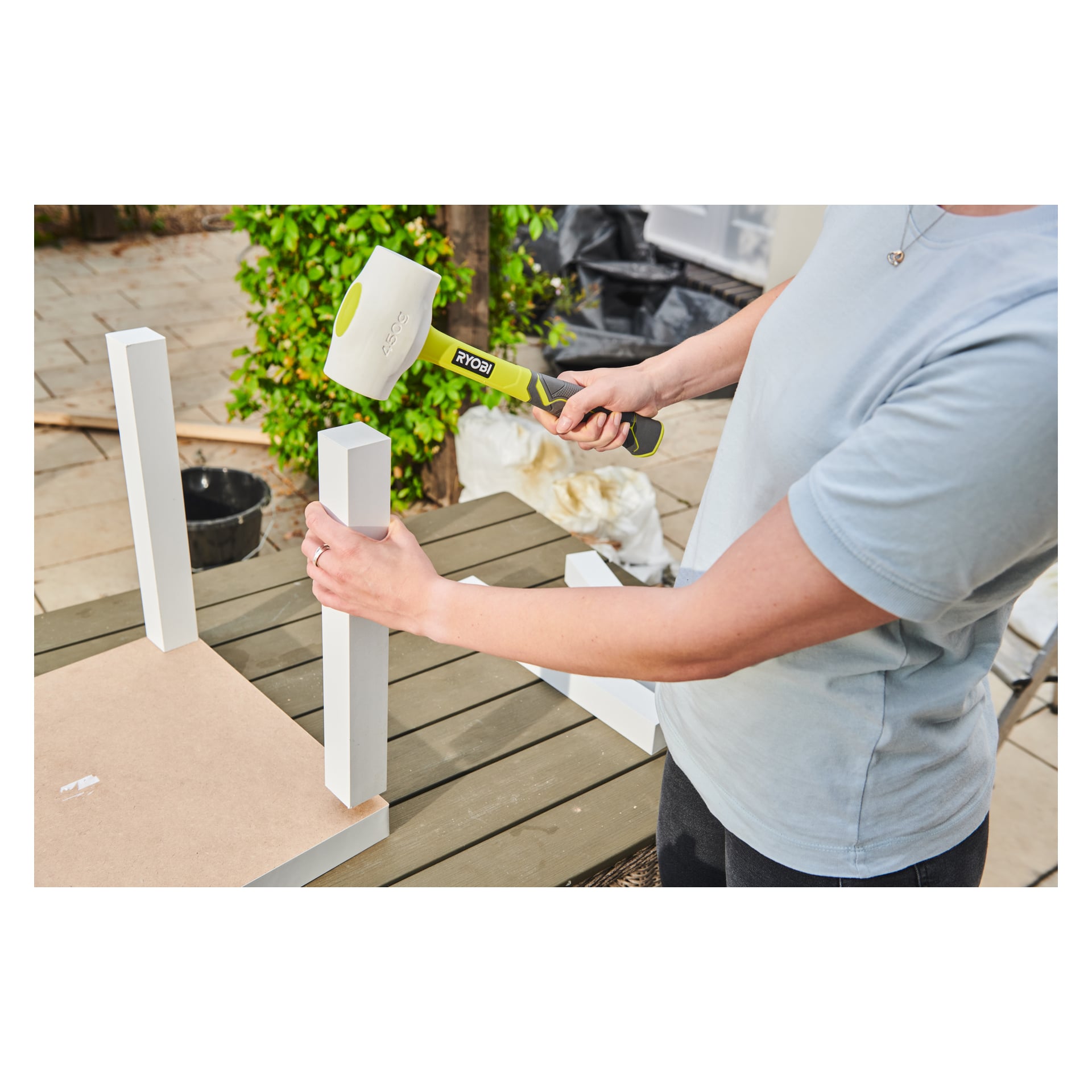 Någon använder en Ryobi-gummihammare RHHM450W för att fästa ett bordsben på ett upp-och-nervänt bord på en träyta utomhus, med olika verktyg, växter och byggmaterial synliga i bakgrunden.