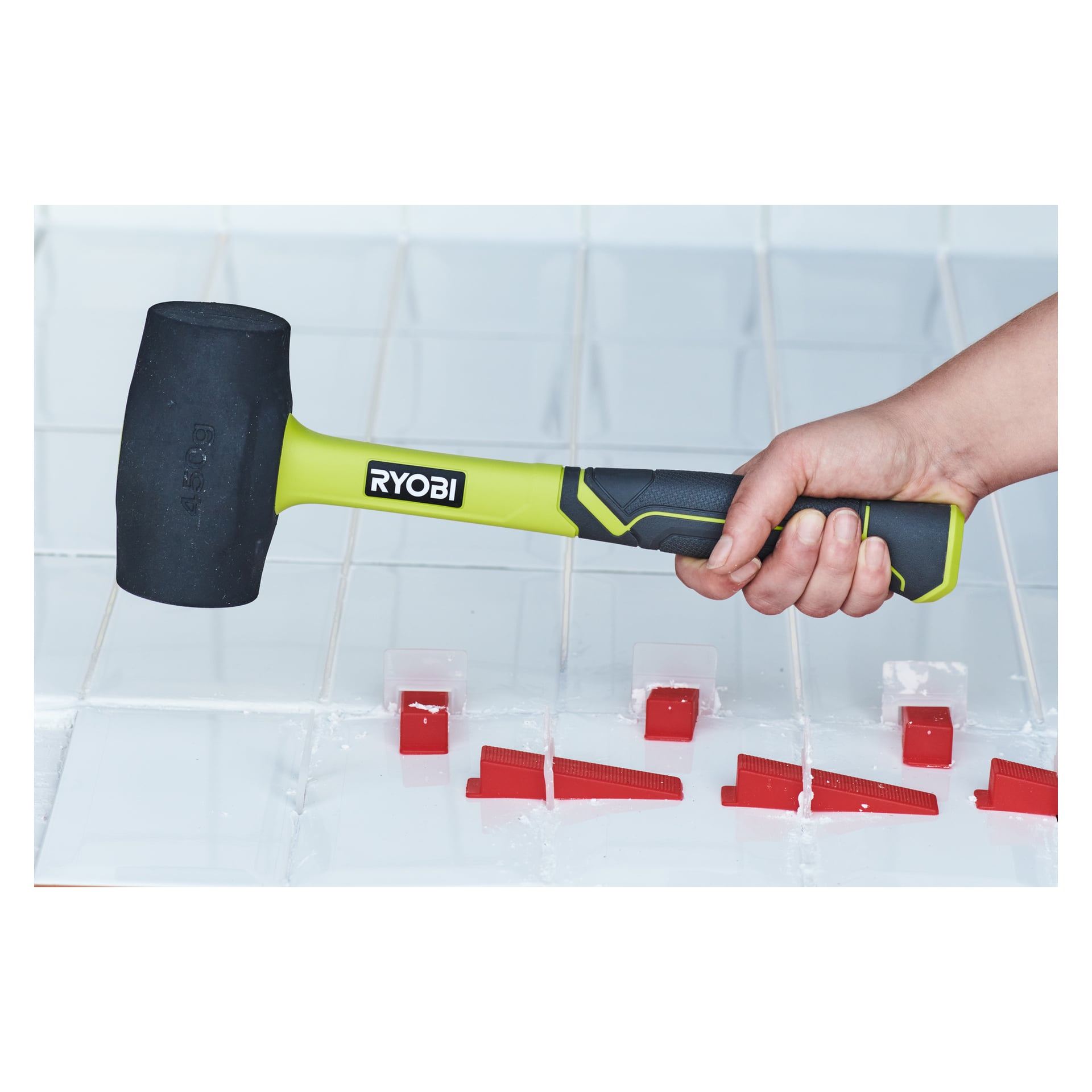 En hand håller en Ryobi Rubber Hammer RHHM450 över vita plattor, där röda plattor är synliga mellan plattorna, vilket indikerar aktiv kakelinstallation.