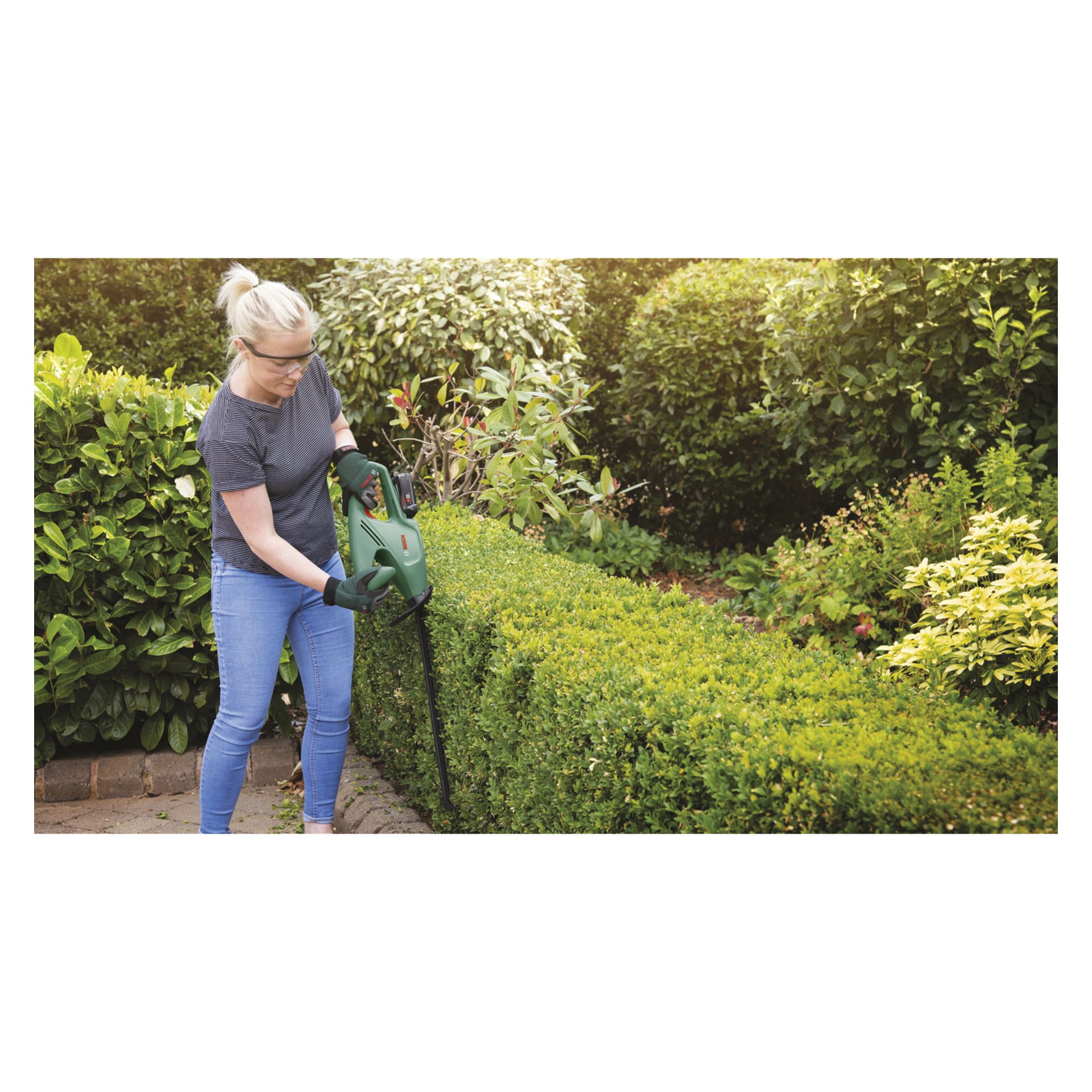 Med skyddsglasögon och handskar trimmar man en frodig grön häck med en Bosch Power 4 All EasyHedgeCut 18-45 batteridriven häcksax i en trädgård fylld med olika buskar och växter i bakgrunden.